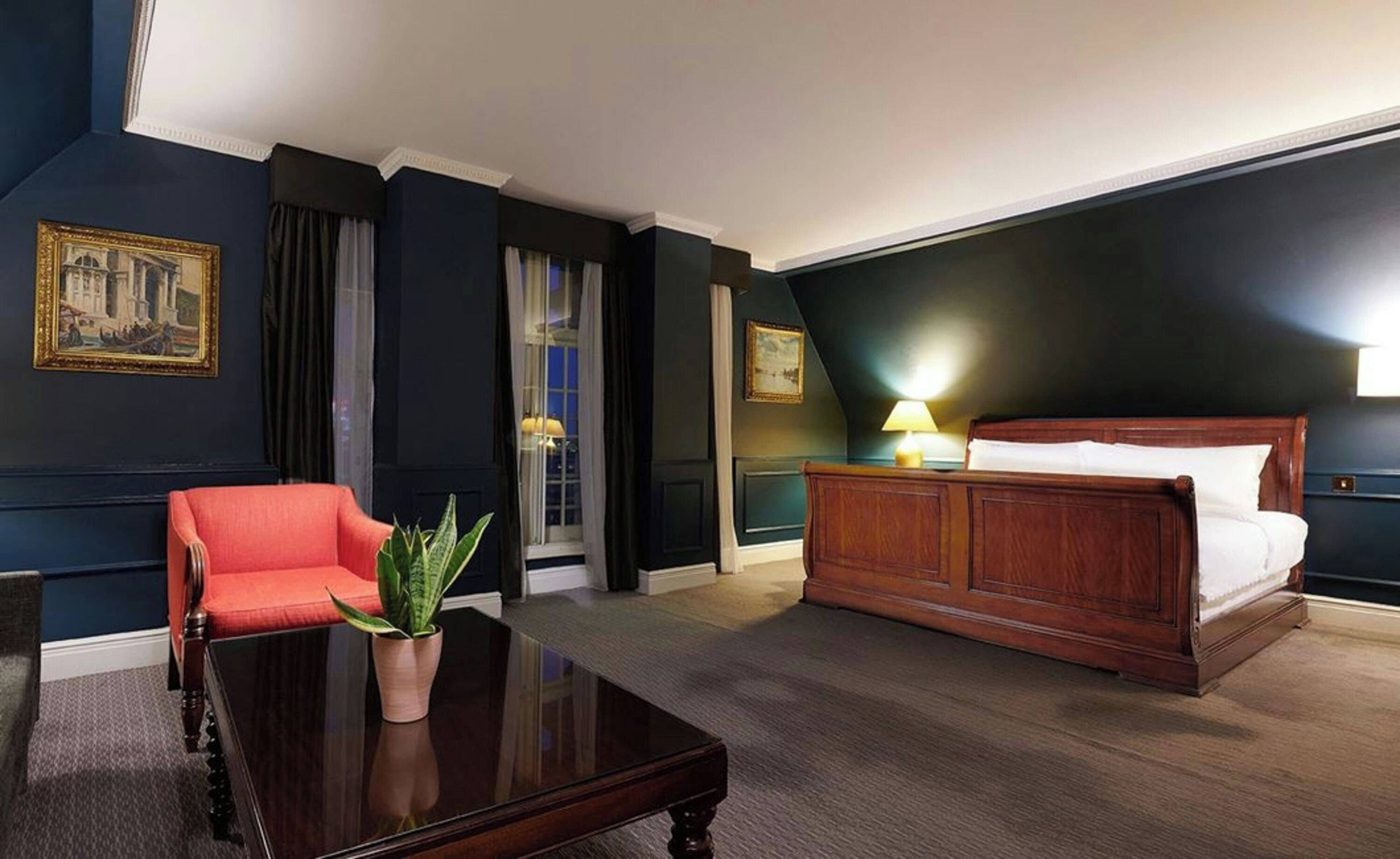 A hotel room with a large bed, a red armchair, a wooden coffee table, and a potted plant.