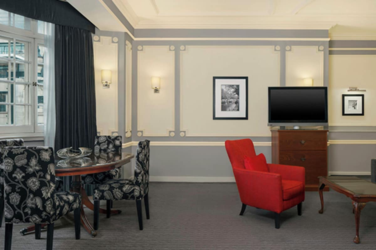 A hotel room with a dining area, a red armchair, a sofa, a television, and framed pictures on the wall.