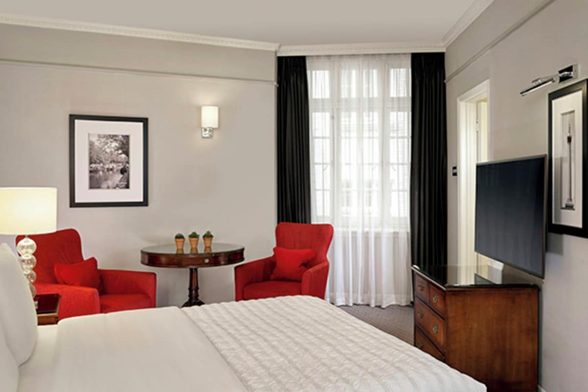 A hotel room with a bed, two red chairs, a small table, a TV, and a framed picture on the wall.