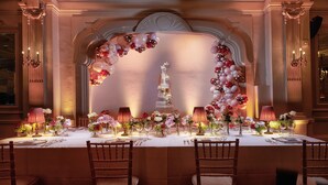 A formal dining room with a long table set for a meal, elegant chairs, and a decorative archway with balloons.