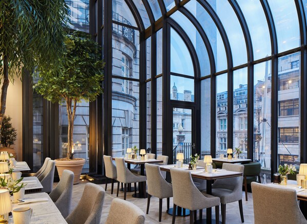 Un ristorante con ampie finestre ad arco, piante da interno e vista sulla città.