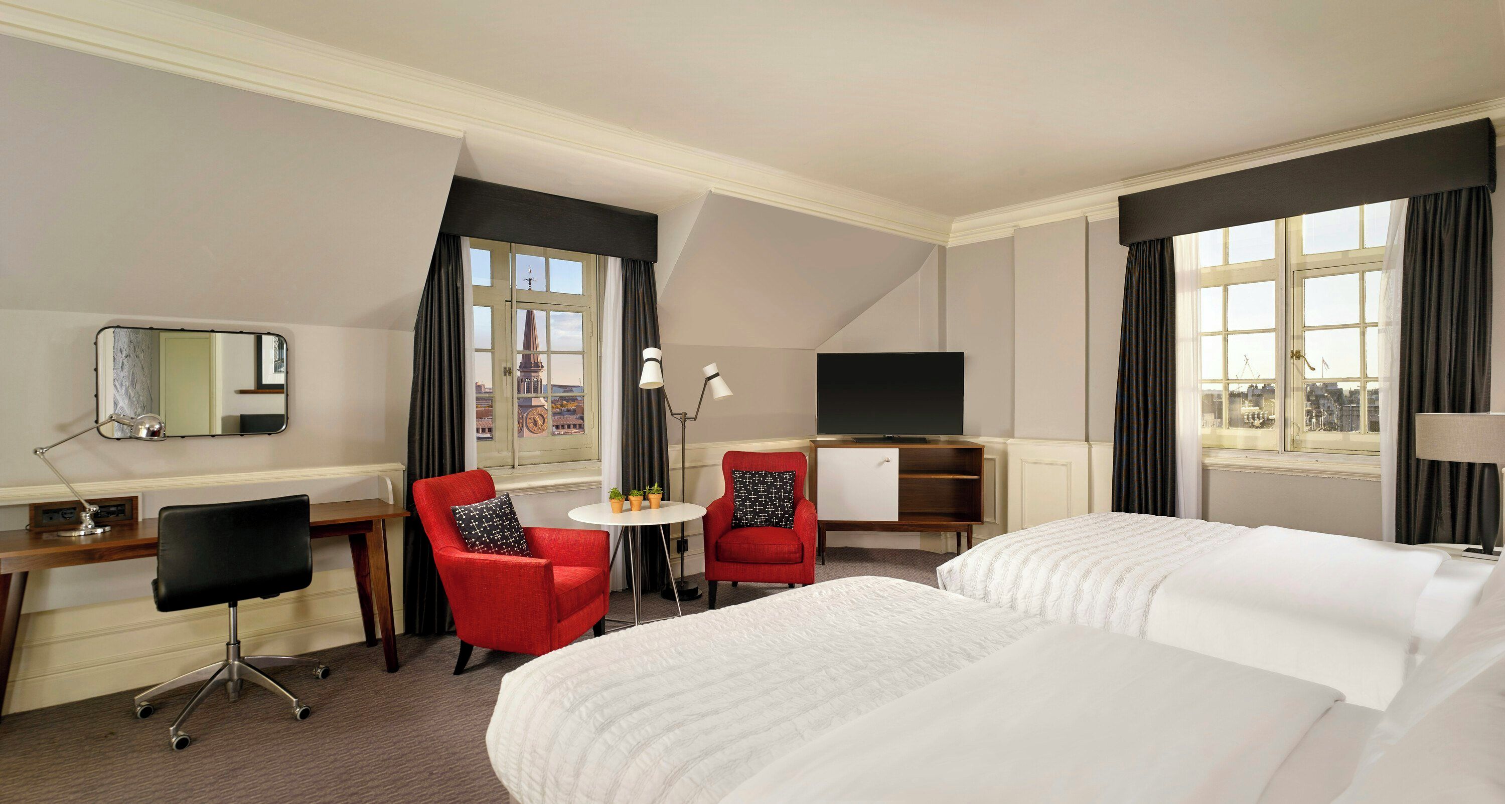 A hotel room with a bed, a desk, two chairs, a TV, and a window with curtains.