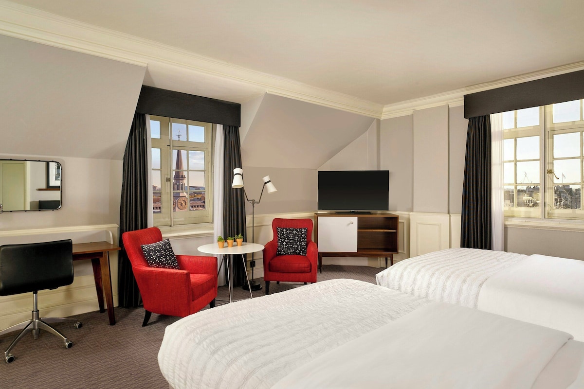 A hotel room with a bed, two red chairs, a desk with a computer, a TV, and a window with curtains.