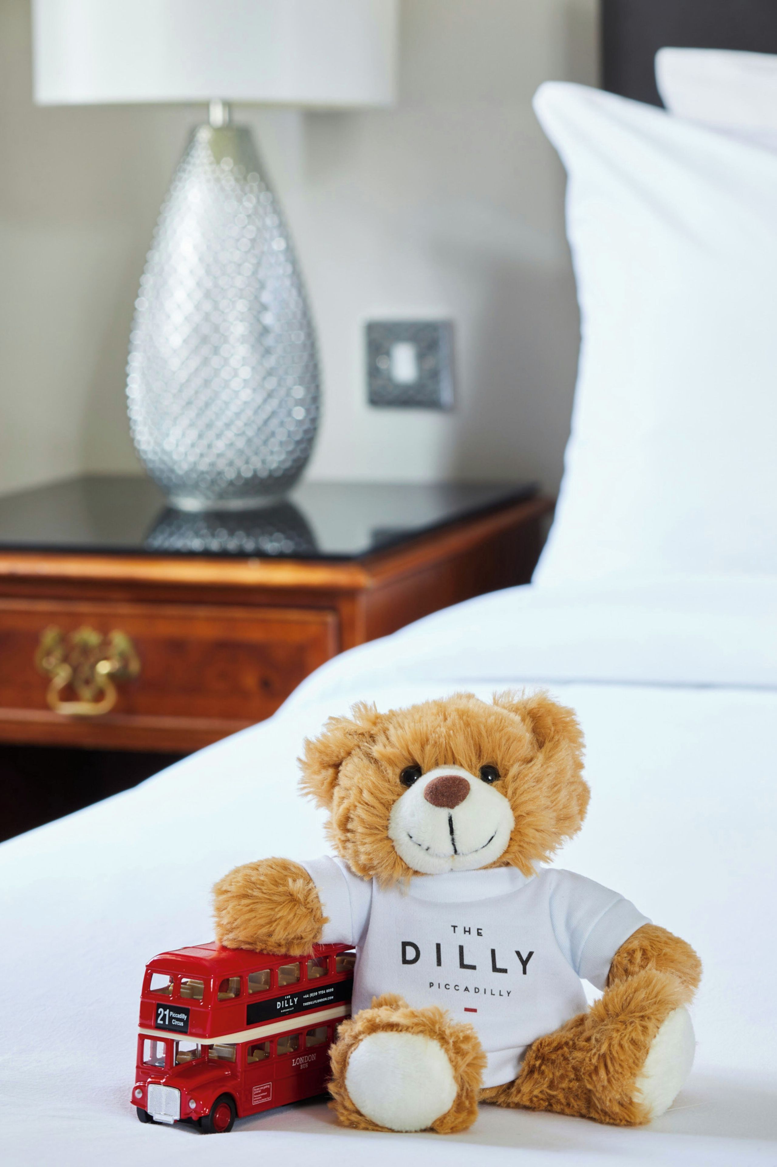 A teddy bear sitting on a bed with a toy red double-decker bus beside it.