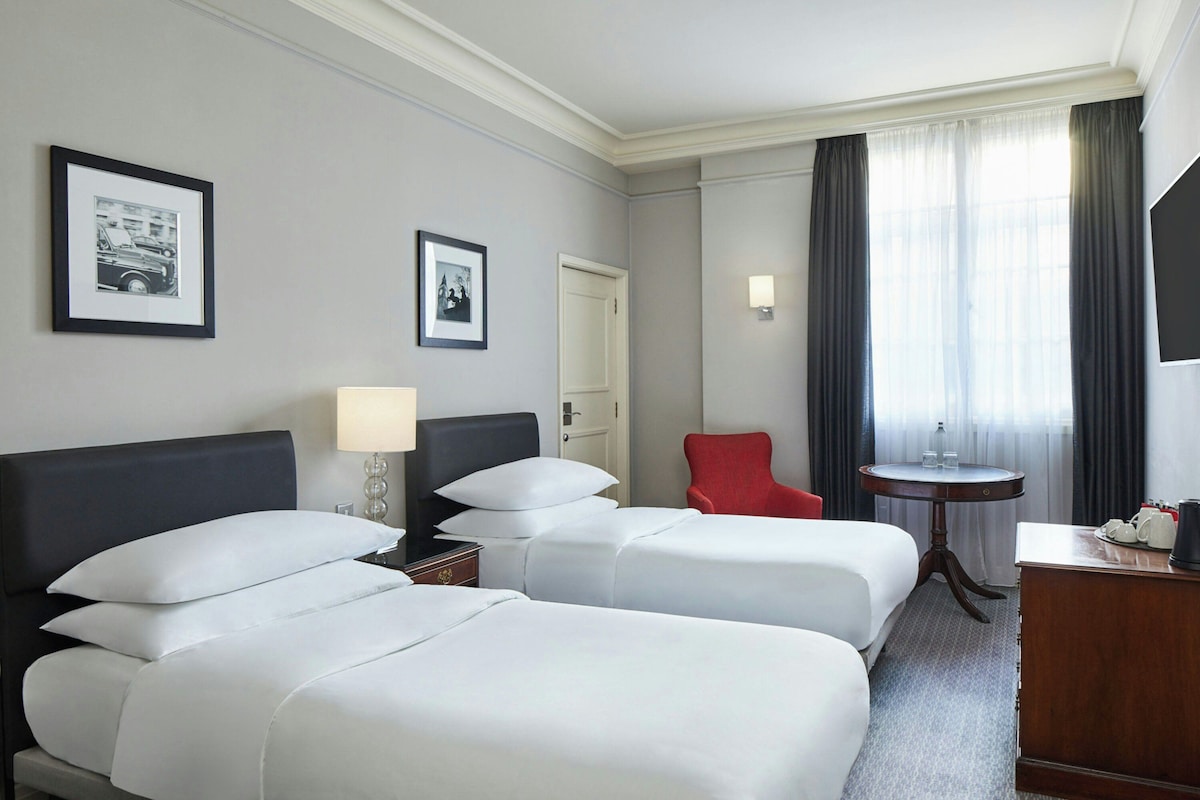 A hotel room with two beds, a red chair, a small table, and a lamp.