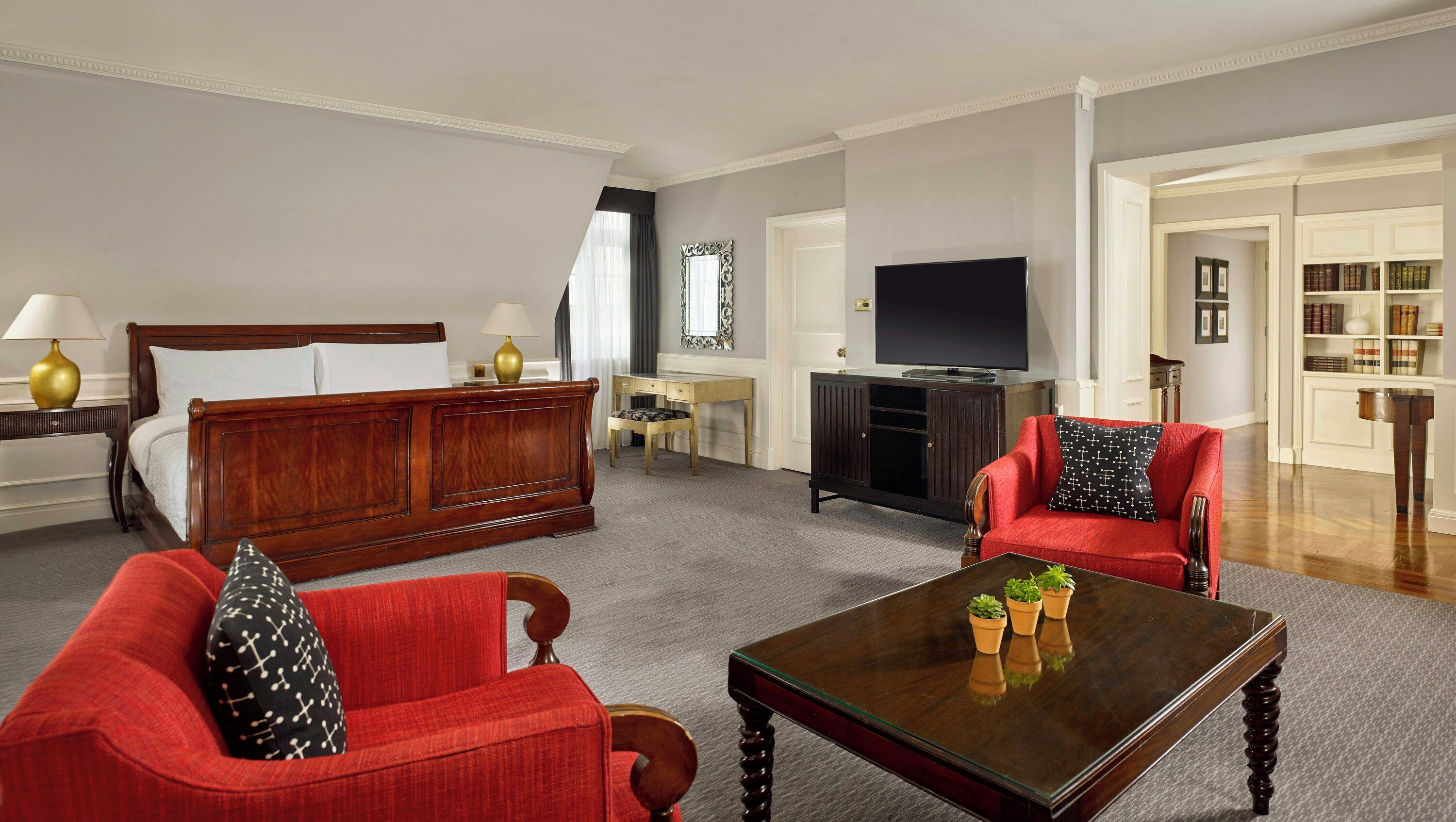 A hotel room with a bed, a red armchair, a sofa, a coffee table, a TV, a bookshelf, and a desk.