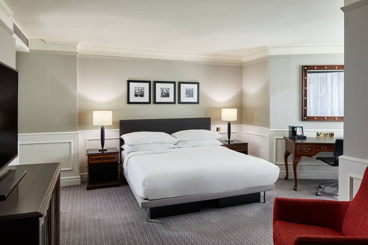 A hotel room with a large bed, two bedside tables with lamps, a desk with a chair, a television, and framed pictures on the wall.