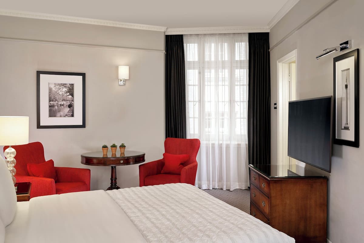 A hotel room with a bed, two red armchairs, a small table with a lamp, a TV, and a framed picture on the wall.
