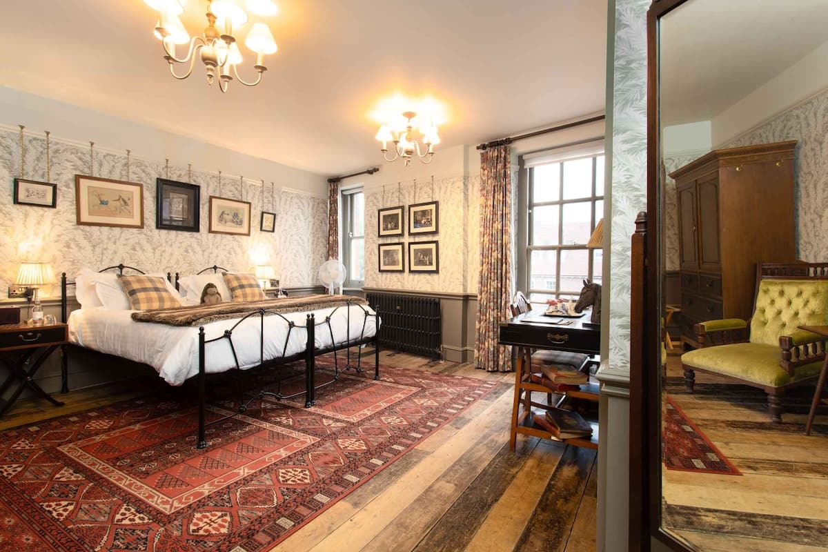 Ein traditionell eingerichtetes Schlafzimmer mit einem Bett, einem Schreibtisch und einem Sessel. An der Wand hängen gerahmte Bilder und über dem Bett hängt eine Chandelieber.
