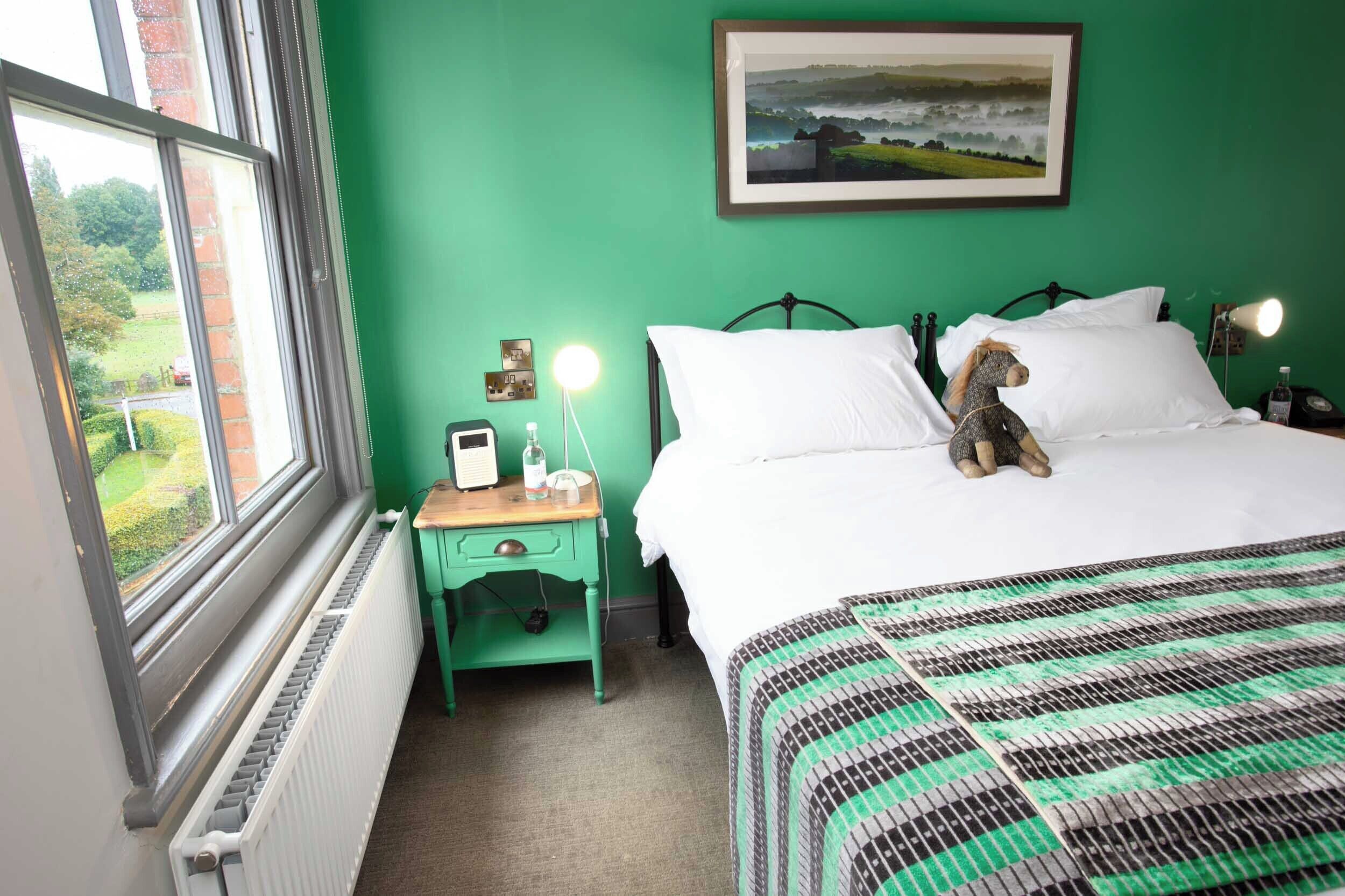 A bedroom with a green wall, a bed with a striped bedspread, a nightstand with a lamp, a window with a view of greenery, and a framed picture above the bed.