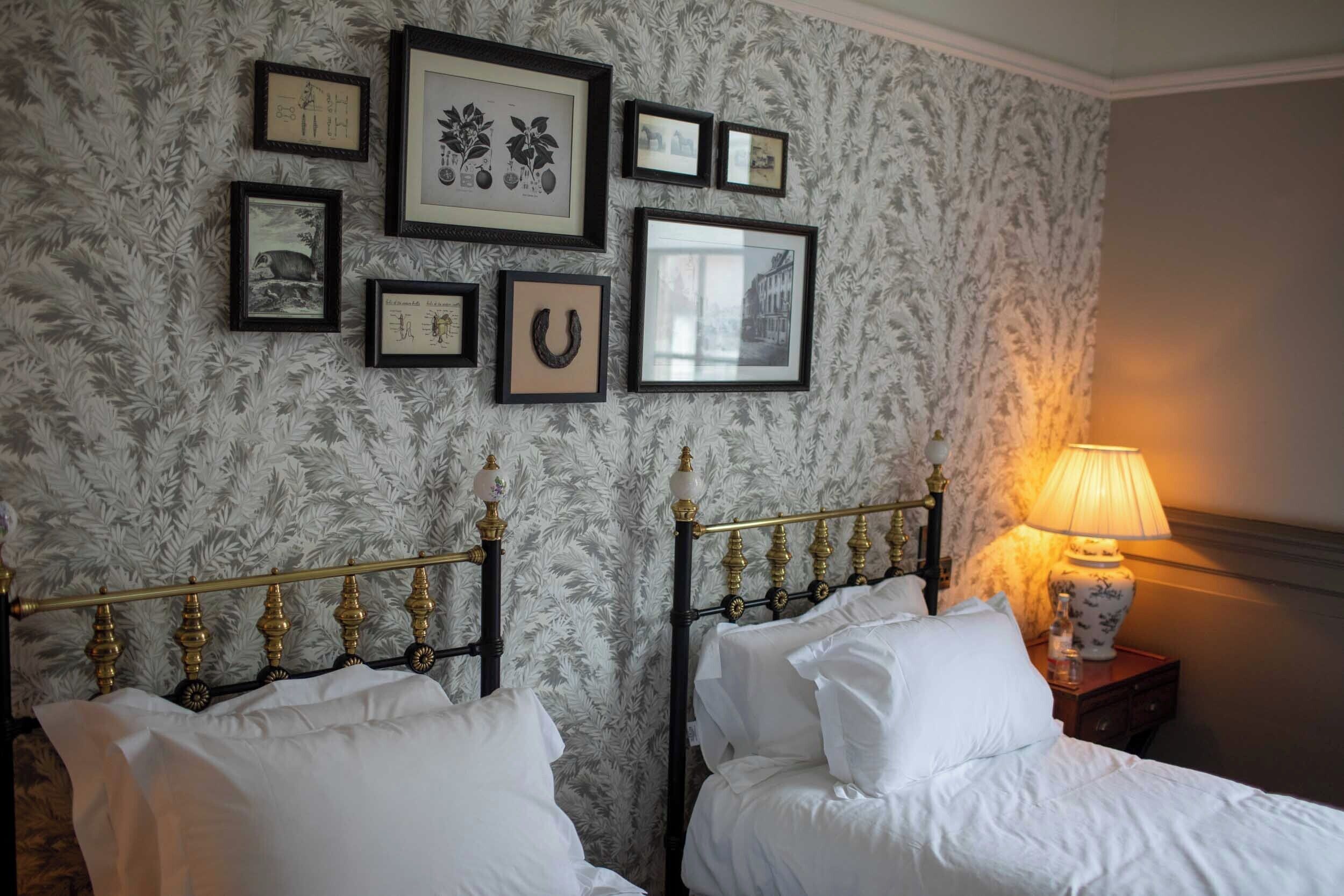 A hotel room with two beds, a patterned wallpaper, and several framed pictures on the wall.