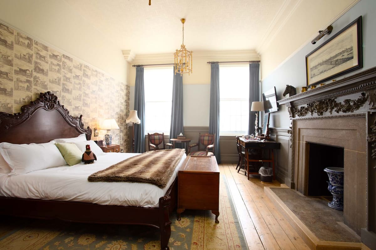 Ein klassisches Hotelzimmer mit einem großen Bett, einer Holzkommode, einem Kamin und einem Schreibtisch mit Stühlen.
