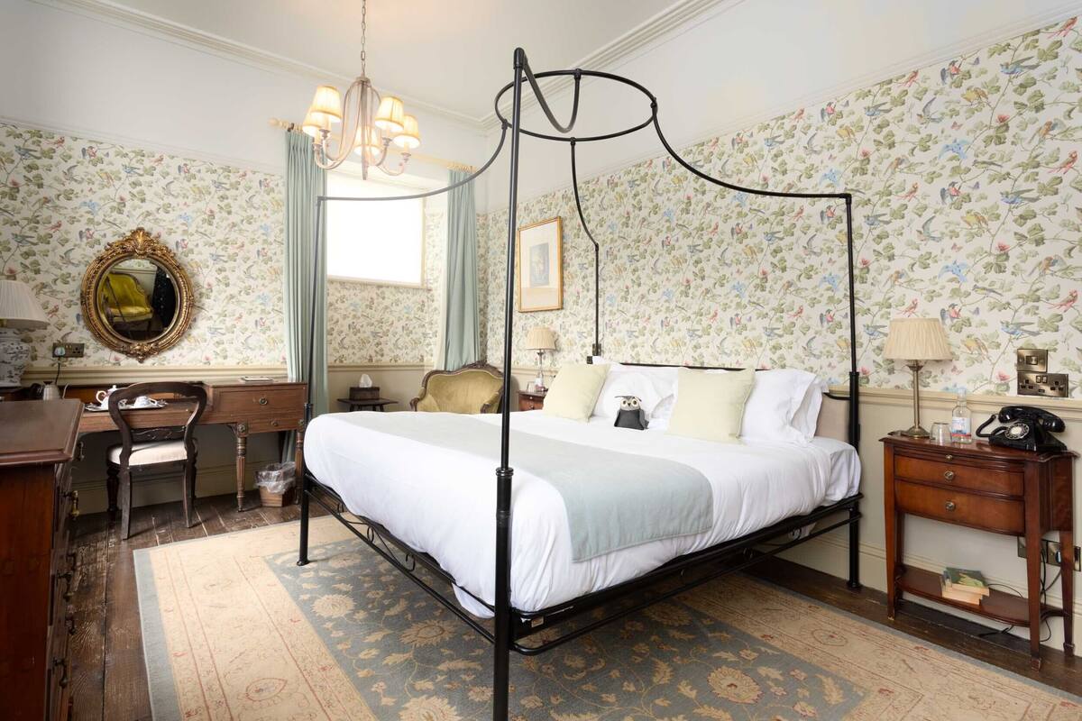 Ein Bett mit Baldachin, ein Schreibtisch mit Stuhl, ein kleiner Nachttisch mit Lampe und ein Telefon im Vintage-Stil.