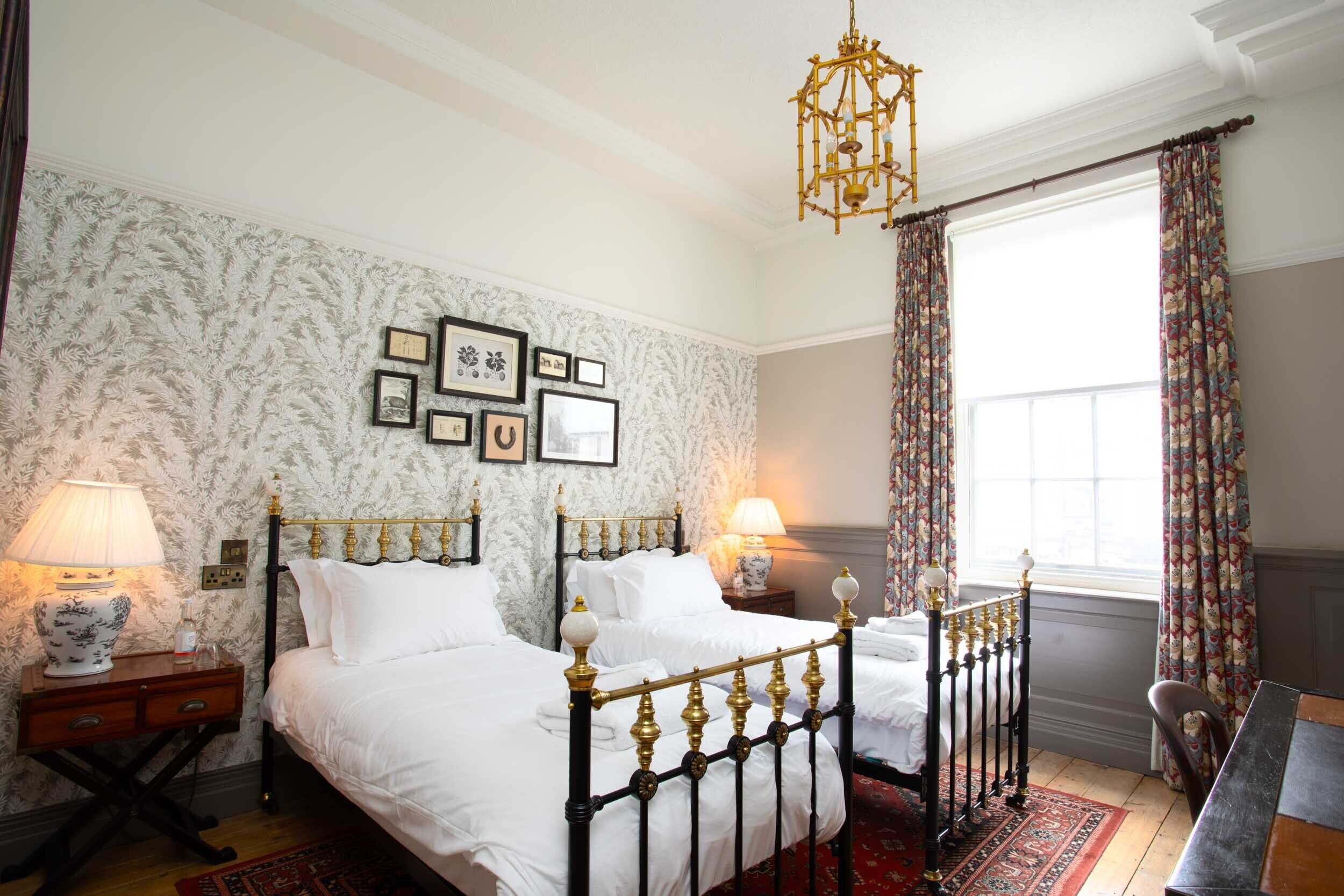 Ein Schlafzimmer mit zwei Betten, einer Chandelüster, gerahmten Bildern an der Wand und einem Fenster mit Vorhängen.