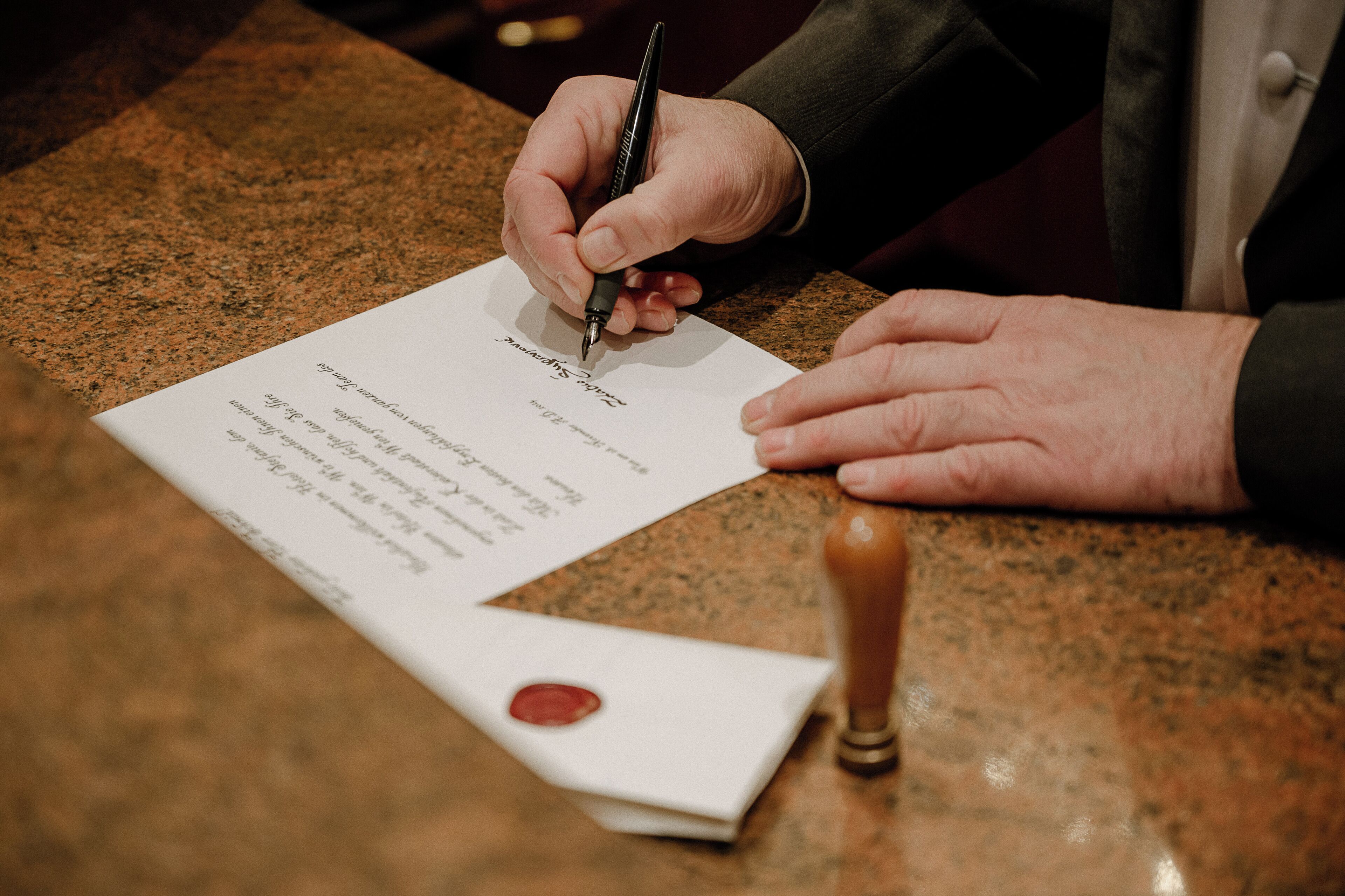 Una persona firmando un documento sobre una mesa de granito, con un sello de cera cerca.
