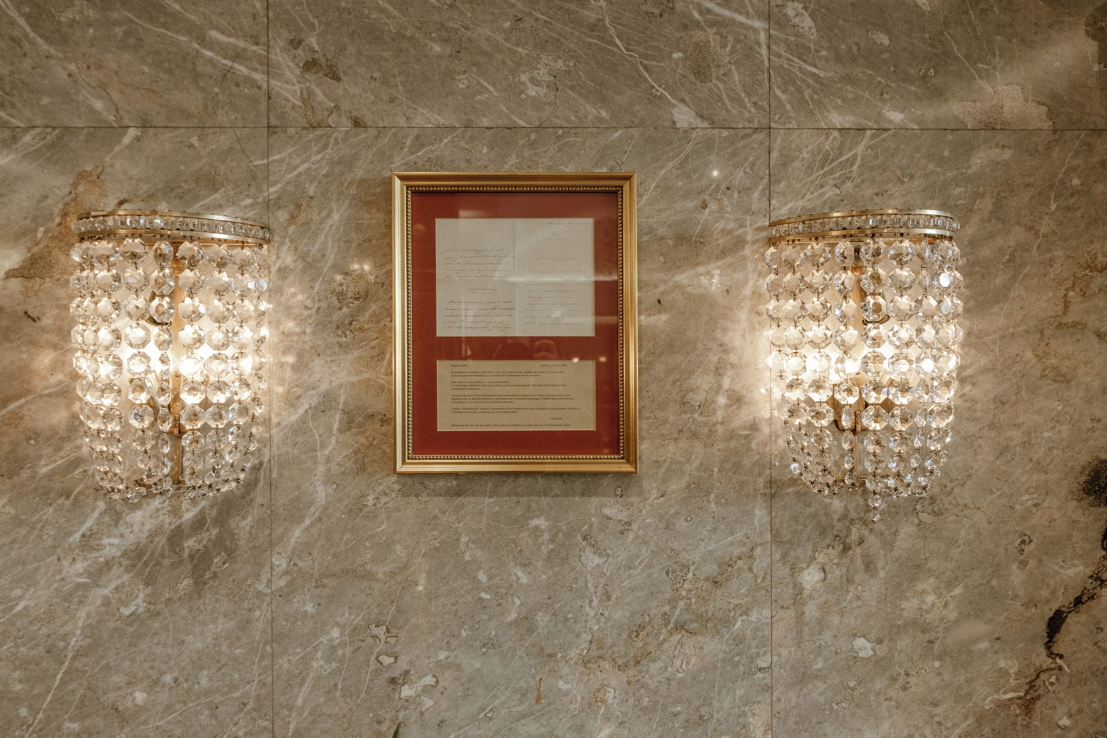 A framed document on a marble wall with two crystal chandeliers on either side.