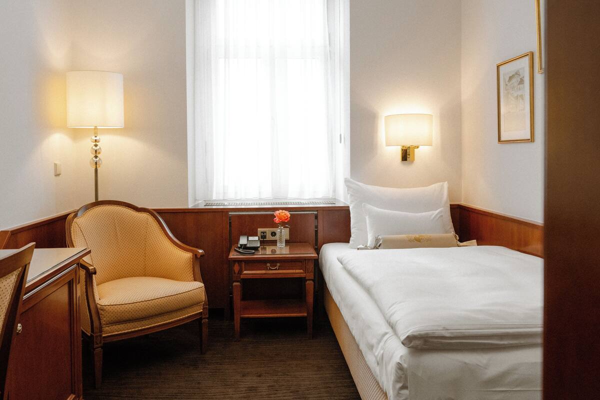 Een hotelkamer met een bed, een stoel, een bureau, een nachtkastje, een lamp en een raam met dunne gordijnen.