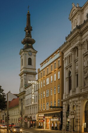 A historic building with a clock tower, Hotel Stefanie, and adjacent shops.