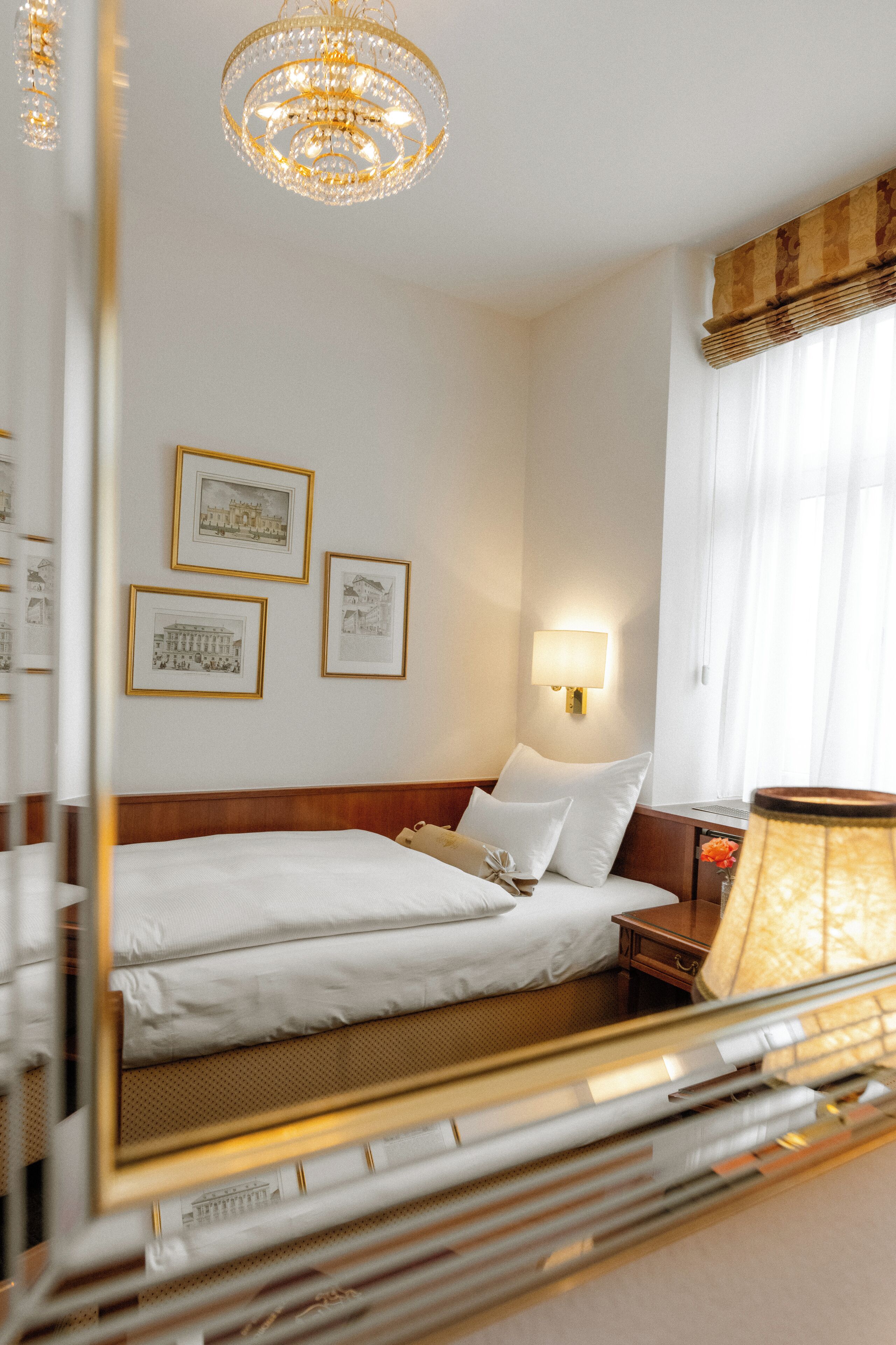 A hotel room with a large bed, a bedside lamp, framed pictures on the wall, and a chandelier.