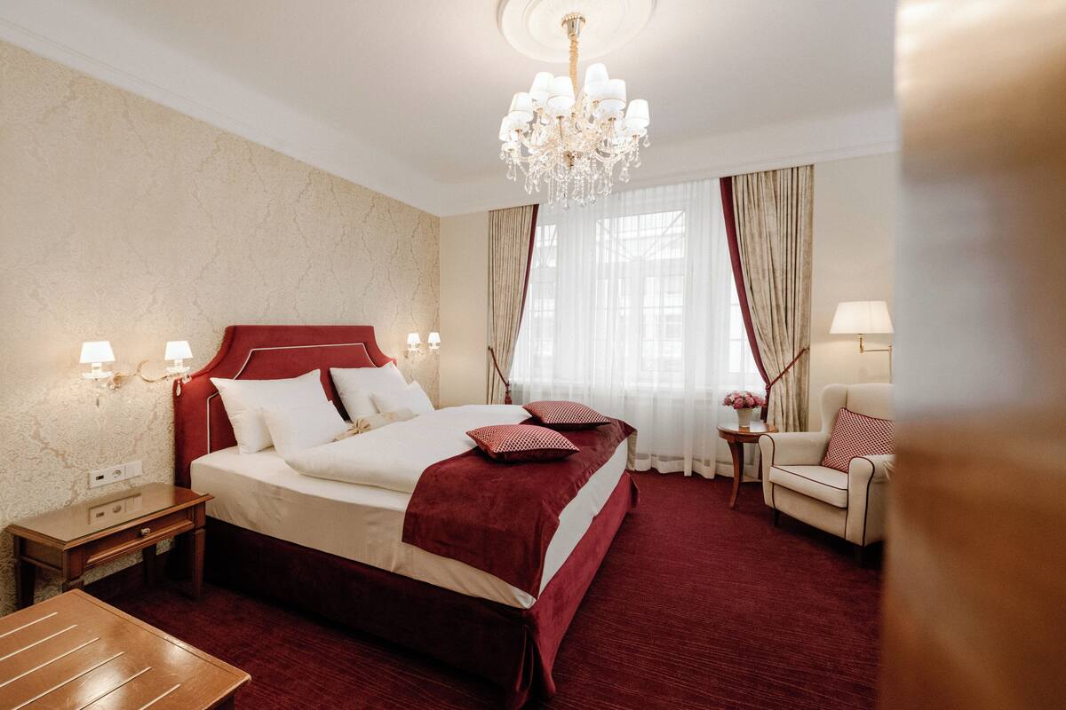 Hotelkamer met een groot bed, een kroonluchter, een zithoek met stoel en tafel, en een raam met gordijnen.