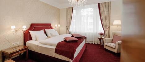 A hotel room with a large bed, a chandelier, a sitting area with a chair and table, and a window with curtains.