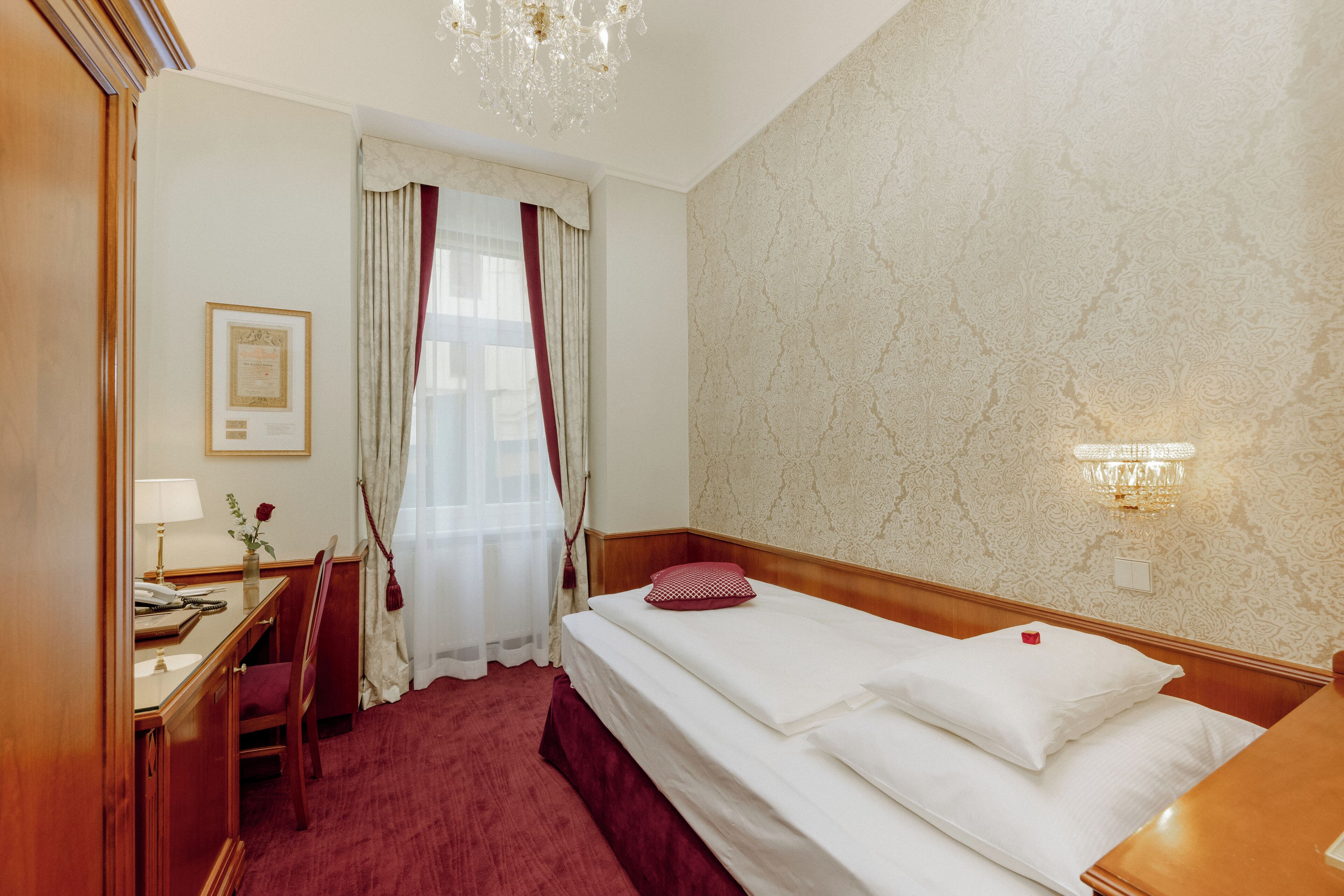 A hotel room with a bed, a desk with a lamp, a chair, a window with curtains, and a chandelier.