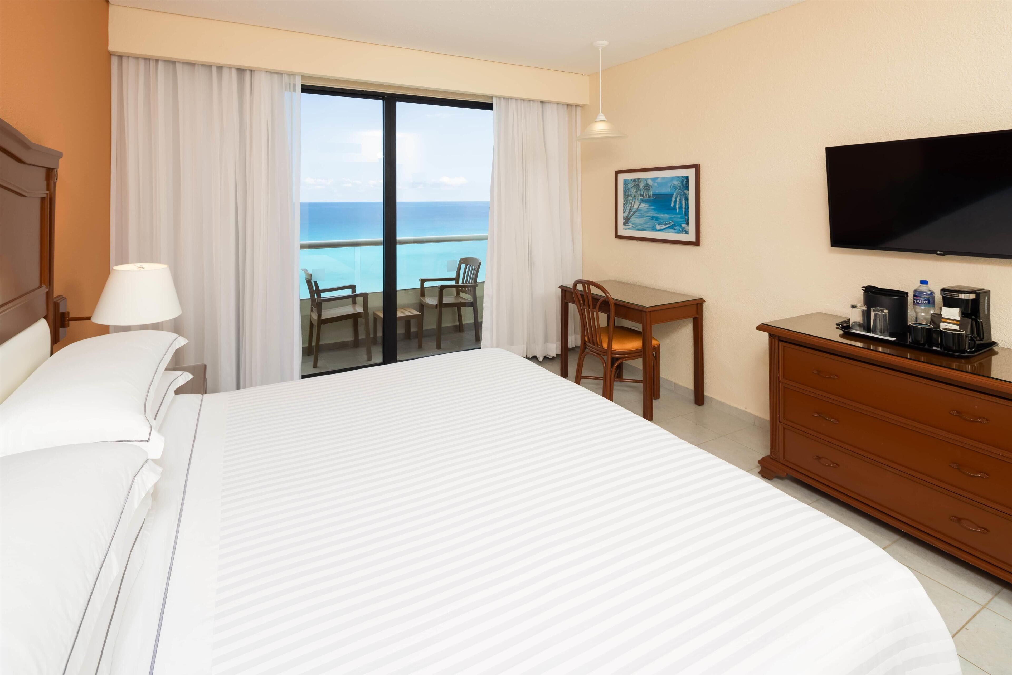 A hotel room with a large bed, a desk, a chair, a television, and a view of the ocean.