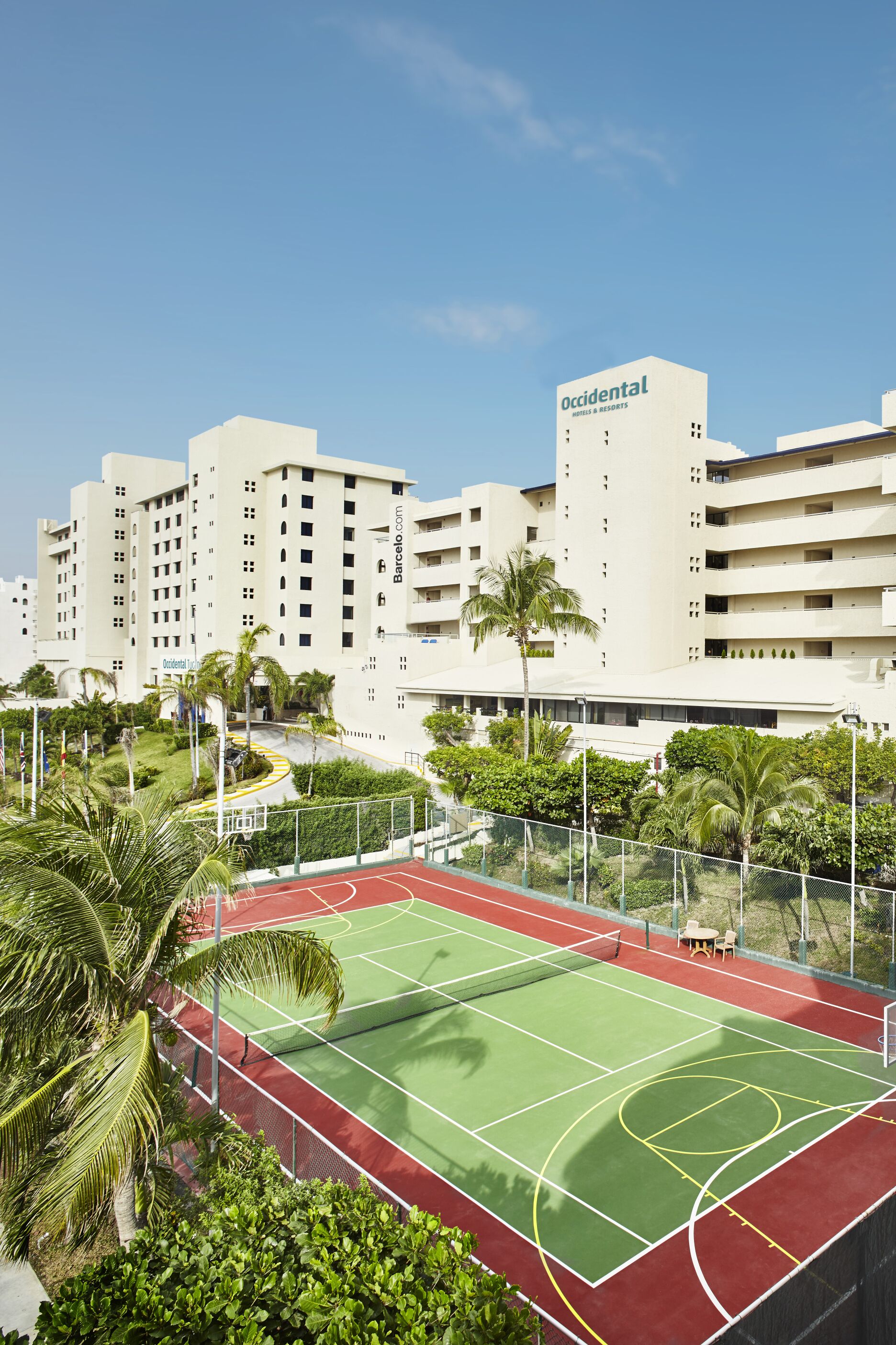 Una cancha de tenis rodeada de vegetación y palmeras, con un edificio de hotel al fondo.