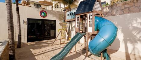 A children's play area with a slide, climbing structures, and a building with a sign.