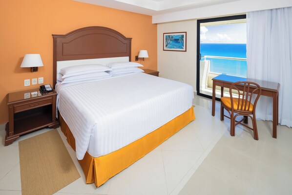 A hotel room with a bed, a desk, a chair, a nightstand, a lamp, and a view of the ocean. - Occidental Tucancún All Inclusive (Cancun)