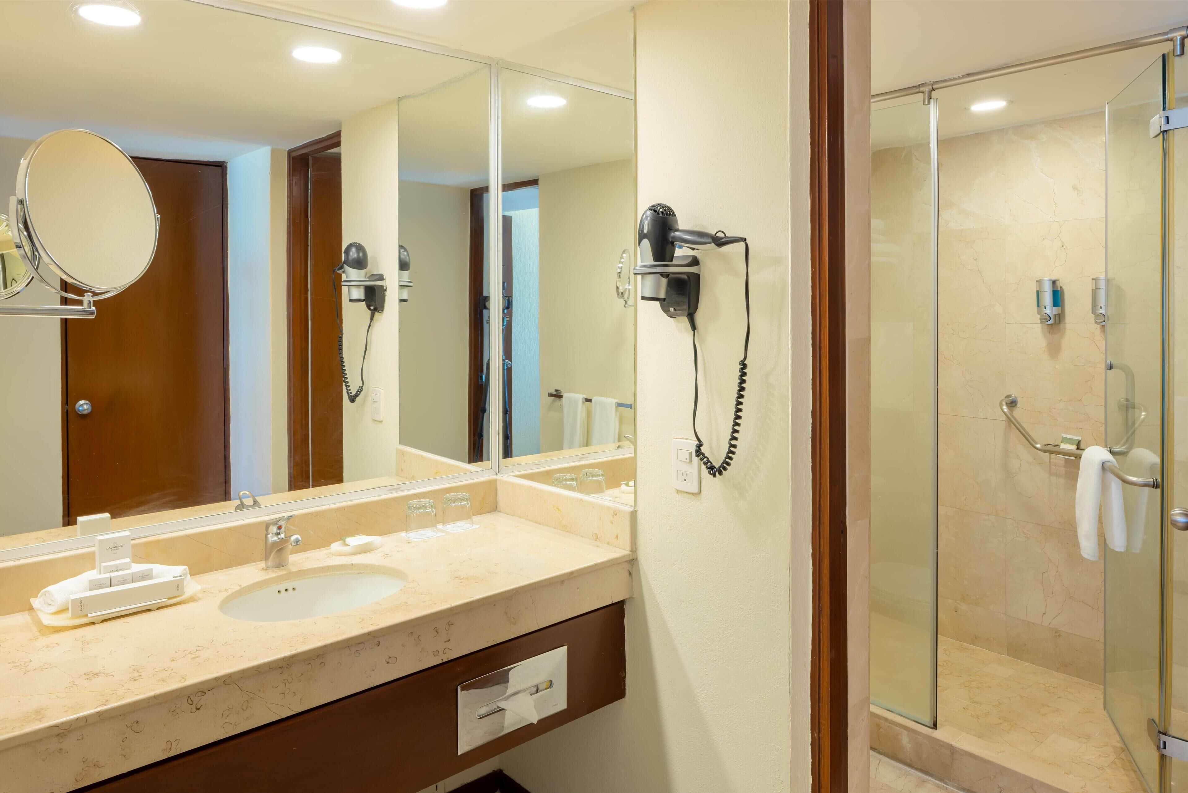 A hotel bathroom with a large mirror, a sink, a hairdryer, and a glass-enclosed shower.