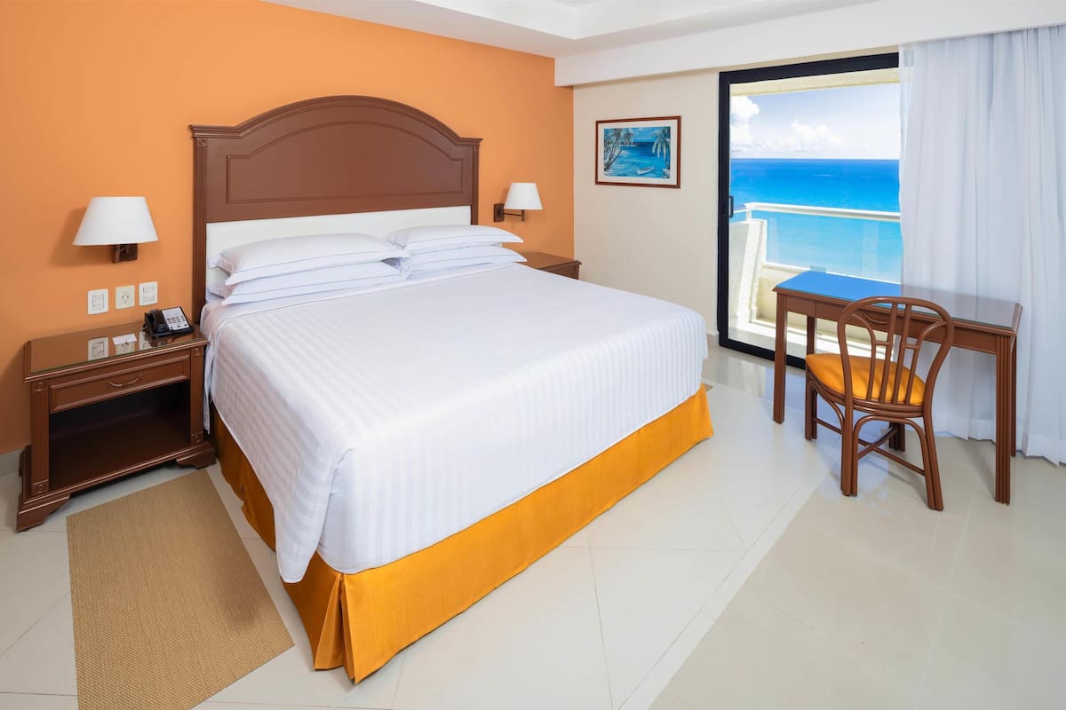 Une chambre d’hôtel avec un grand lit, un bureau avec une chaise, une vue sur l’océan et un tableau au mur.