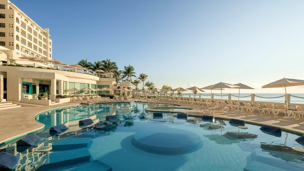A resort pool area with lounge chairs, umbrellas, and a building with multiple floors. - Occidental Tucancún All Inclusive (Cancun)