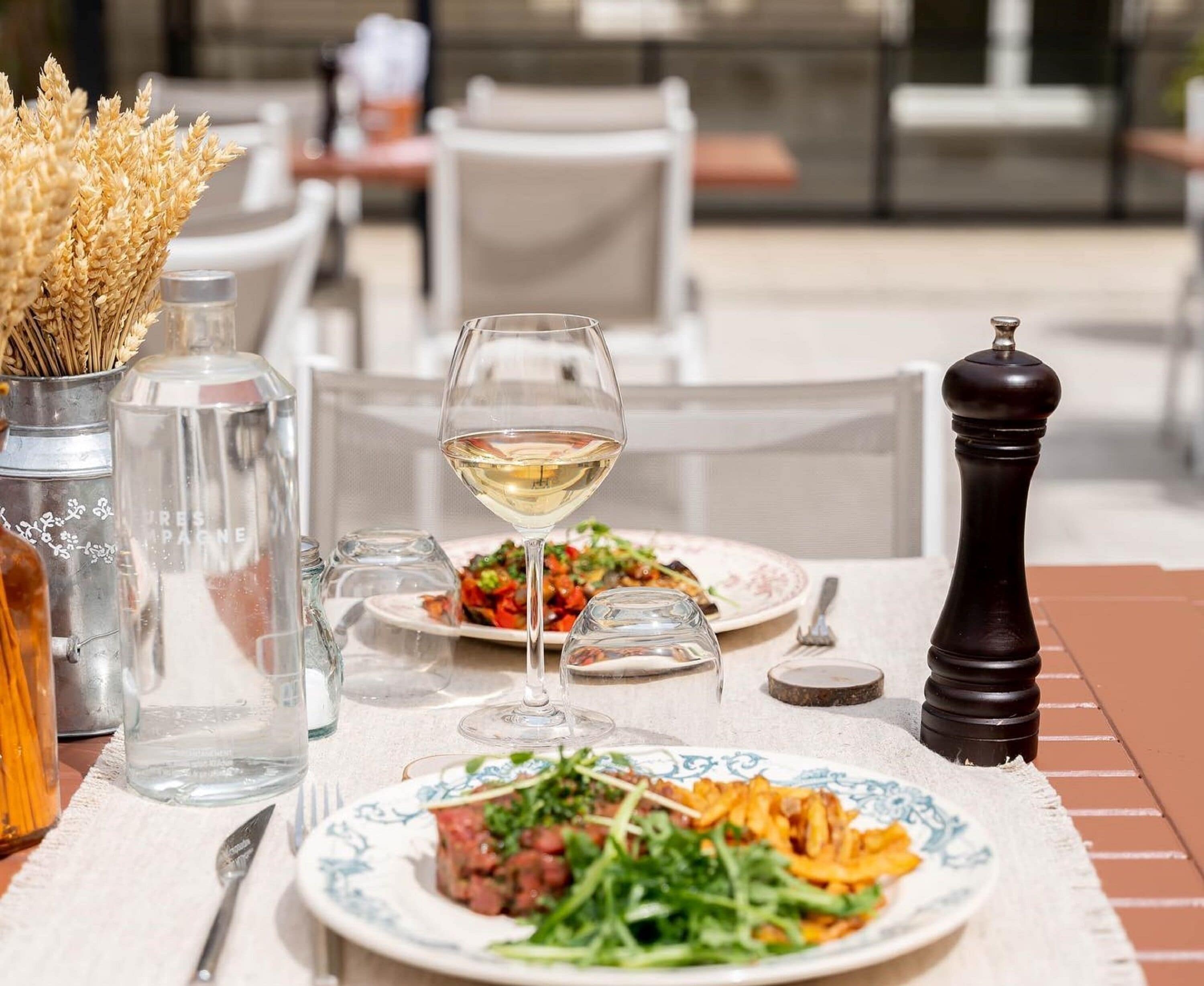 Una mesa dispuesta con platos de comida, una copa de vino blanco, saleros y pimenteros, y un florero con flores secas.