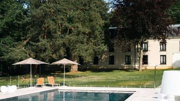 Une piscine entourée d’arbres et d’un bâtiment, avec des chaises longues et des parasols.