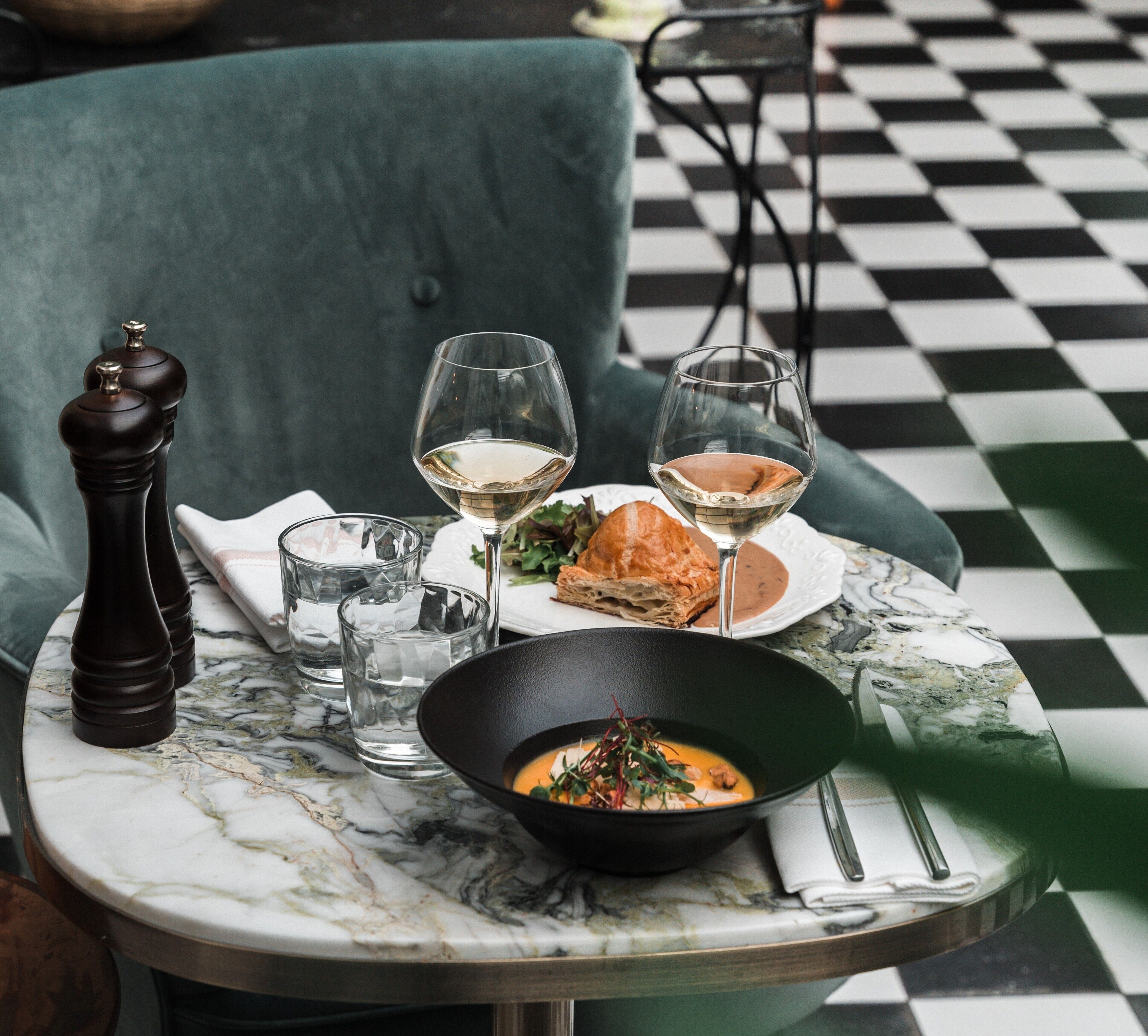 A dining table with a checkered floor, a bowl of soup, two glasses of white wine, a plate with a sandwich, and a pepper grinder.