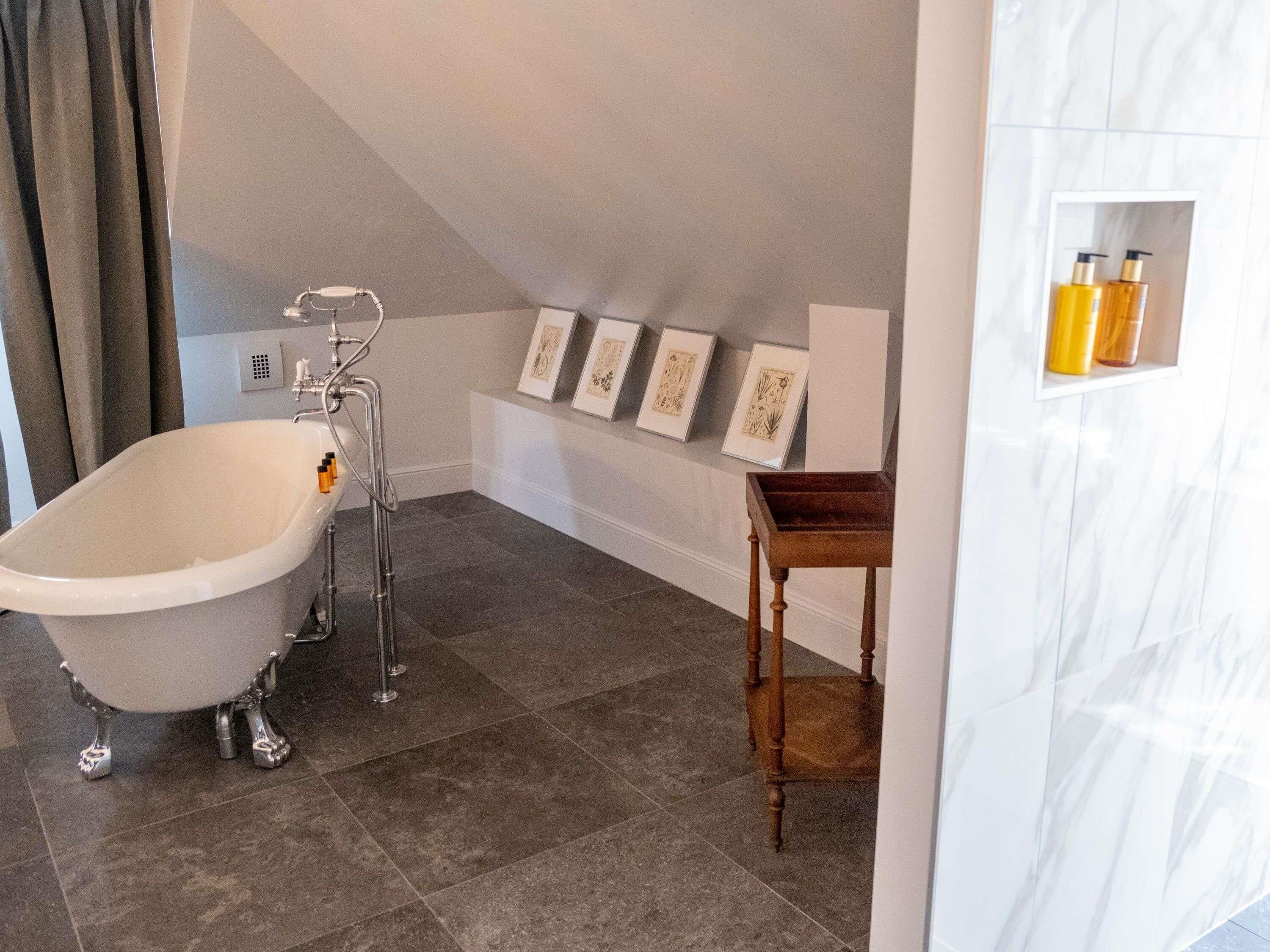 Ein Badezimmer mit einer freistehenden Badewanne, einem hölzernen Beistelltisch und gerahmten Kunstwerken an der Wand.
