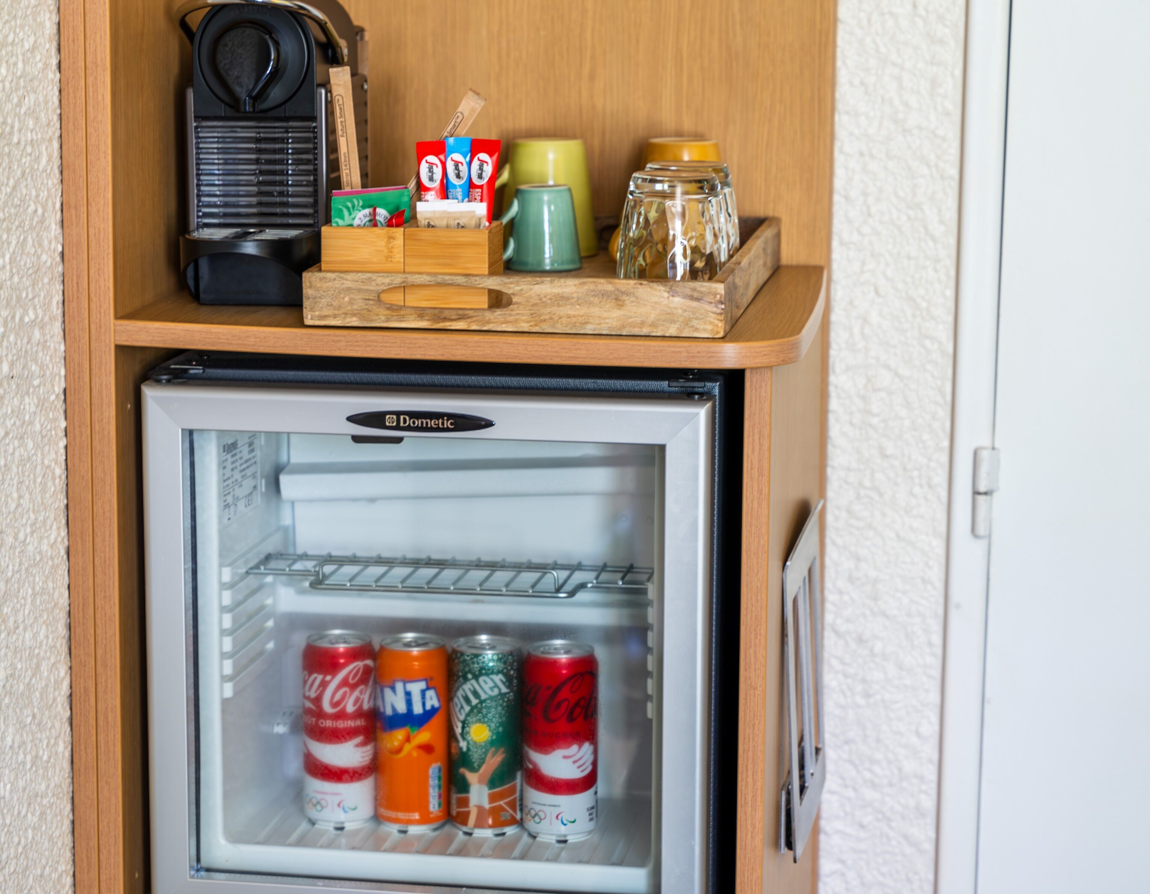 Un mini refrigerador con un estante de madera encima que contiene una cafetera, una taza verde y un frasco. Adentro del refrigerador, hay cuatro latas de refresco.