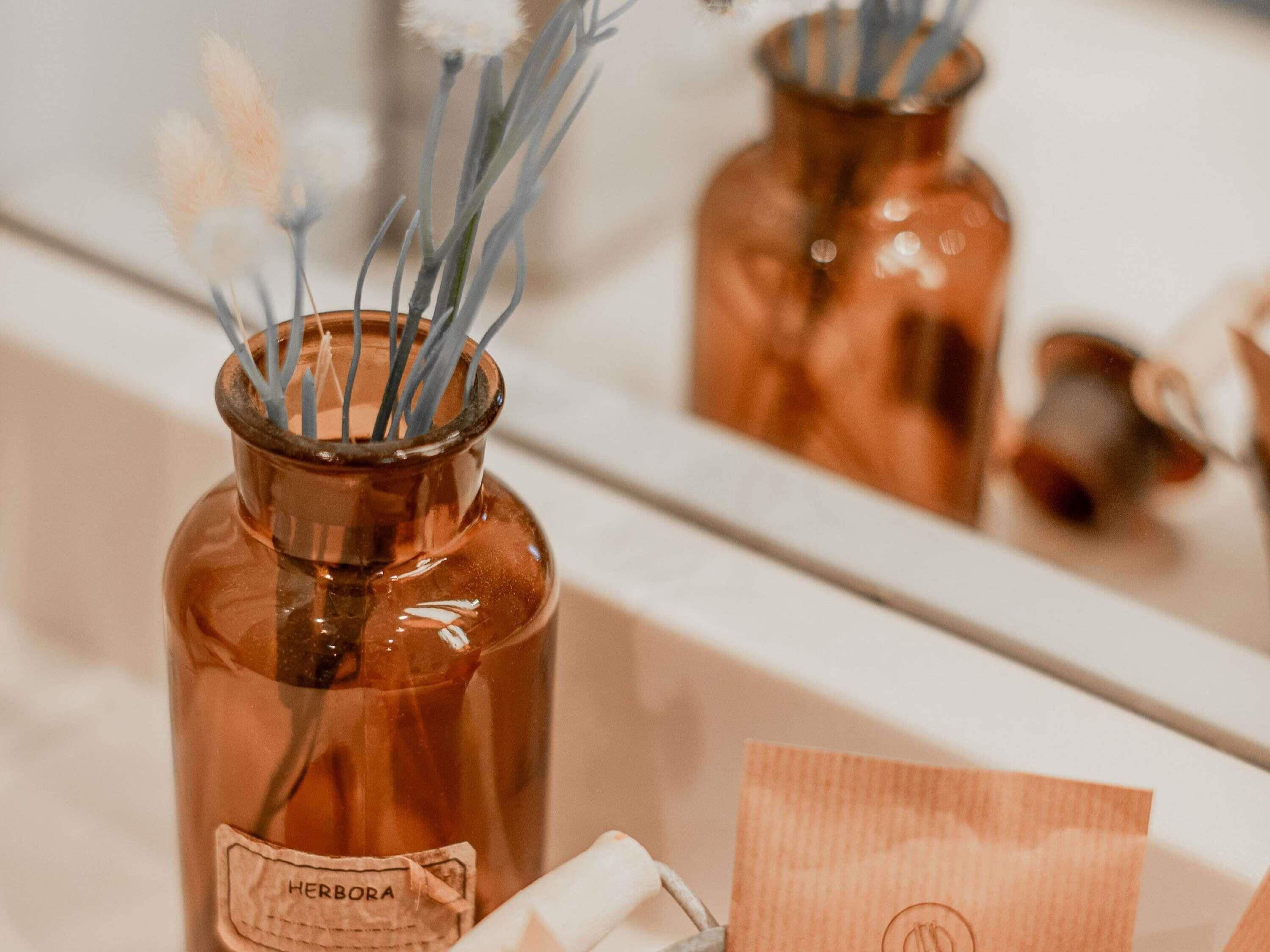 Un baño con un espejo, un florero de color cobre con una etiqueta y un objeto blanco sobre el mostrador.