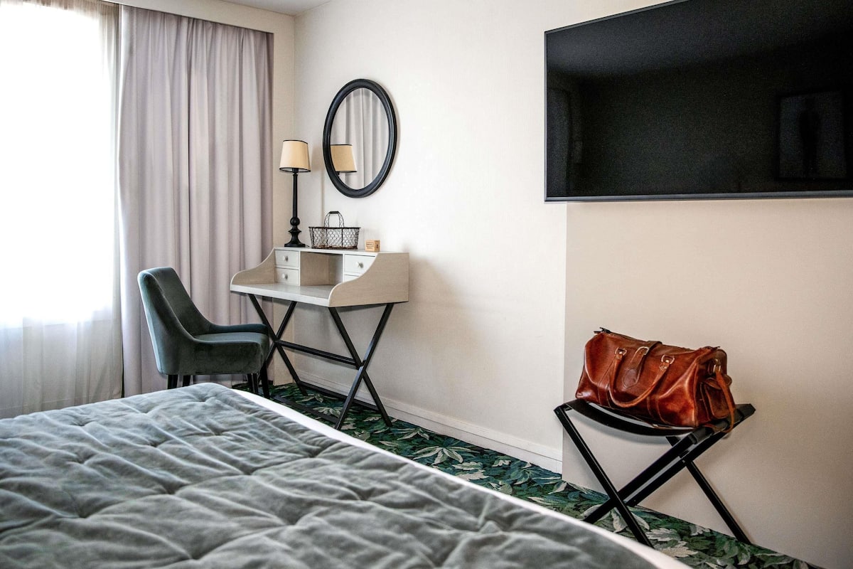 Ein Hotelzimmer mit einem Bett, einem Schreibtisch mit Lampe, einem Sessel, einem kleinen Tisch mit einer Handtasche und einem an der Wand befestigten Fernseher.