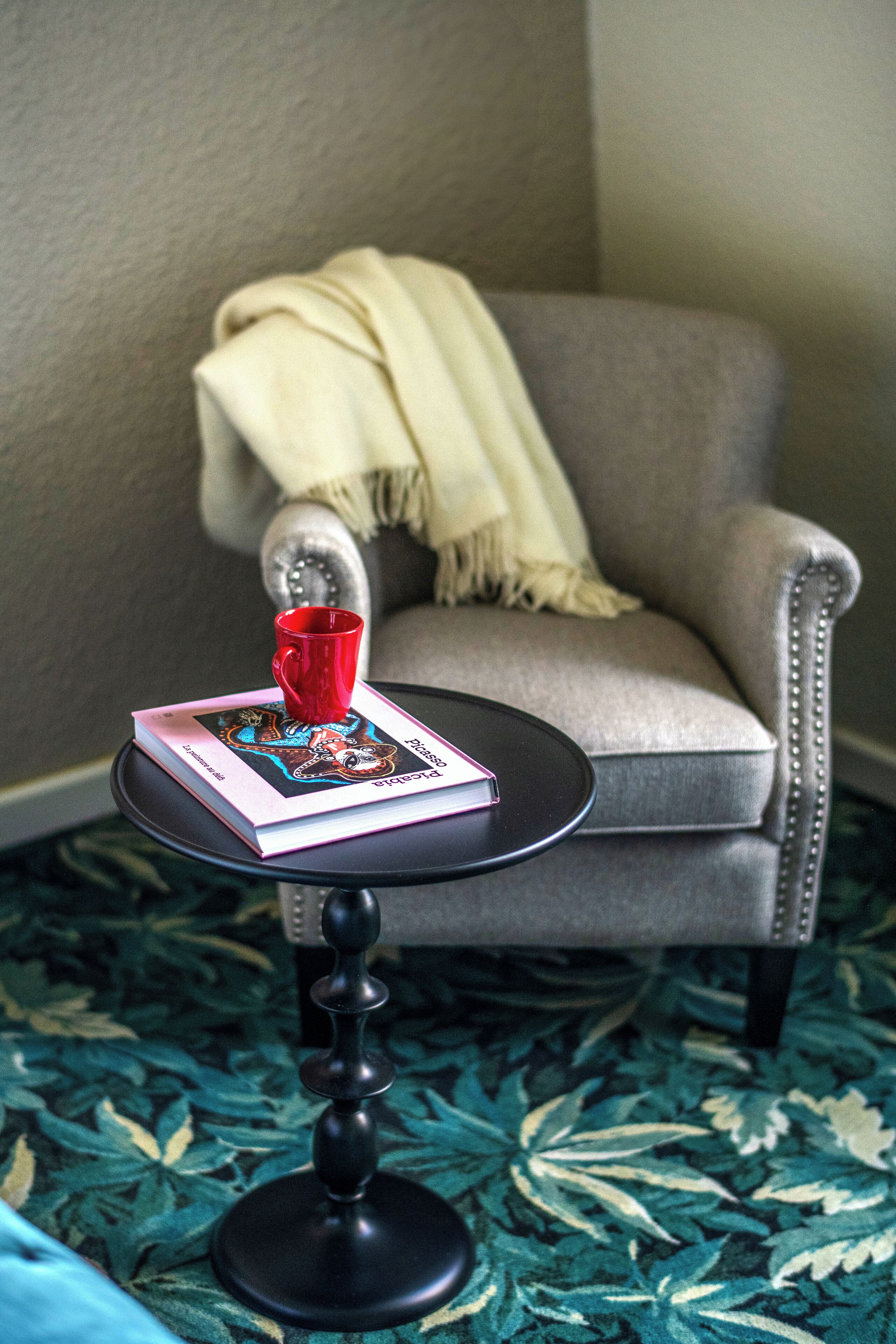 A small round table with a book and a red mug, a grey armchair with a beige throw blanket, and a patterned carpet.