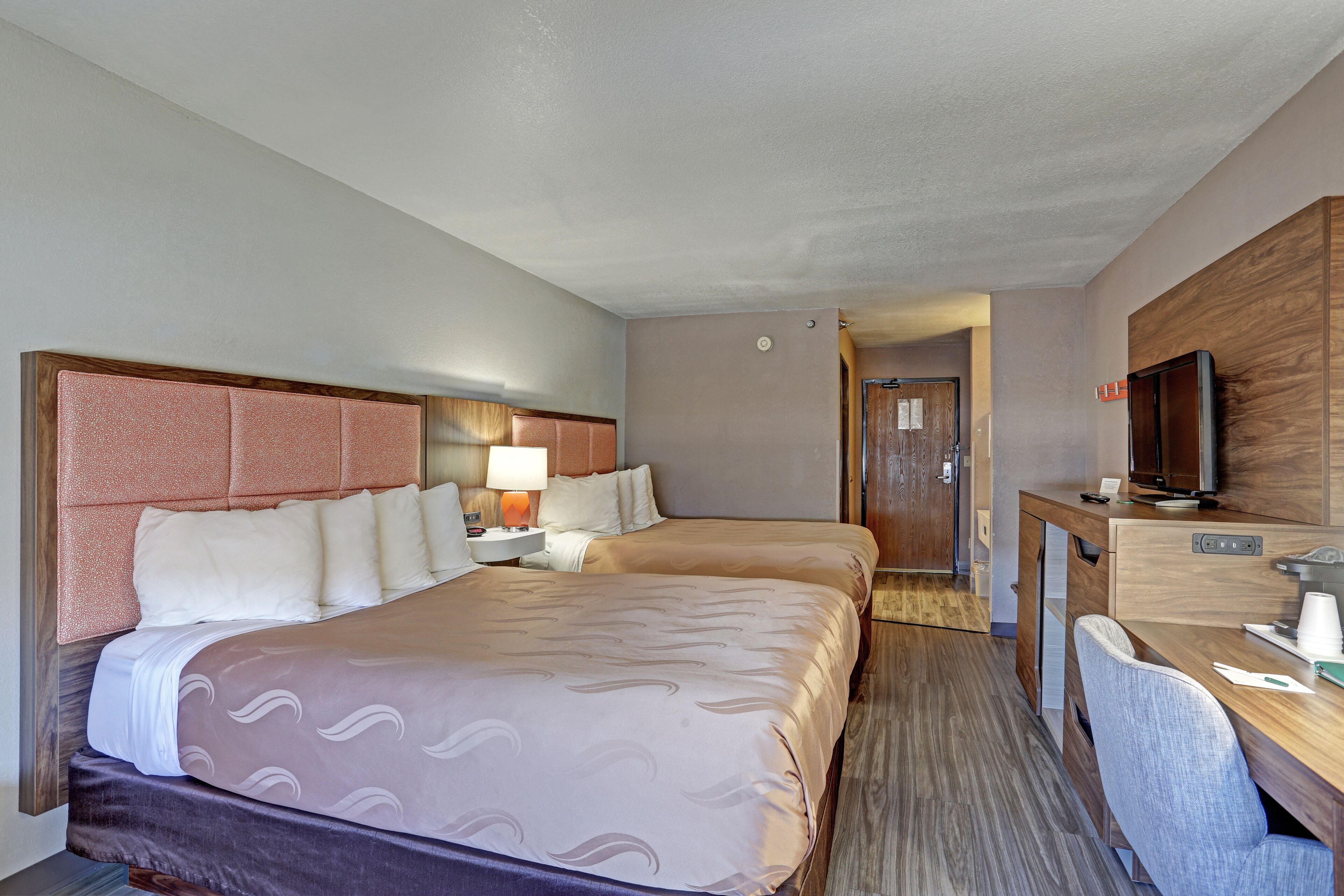 A hotel room with two beds, a desk, a chair, a television, and a nightstand with a lamp.