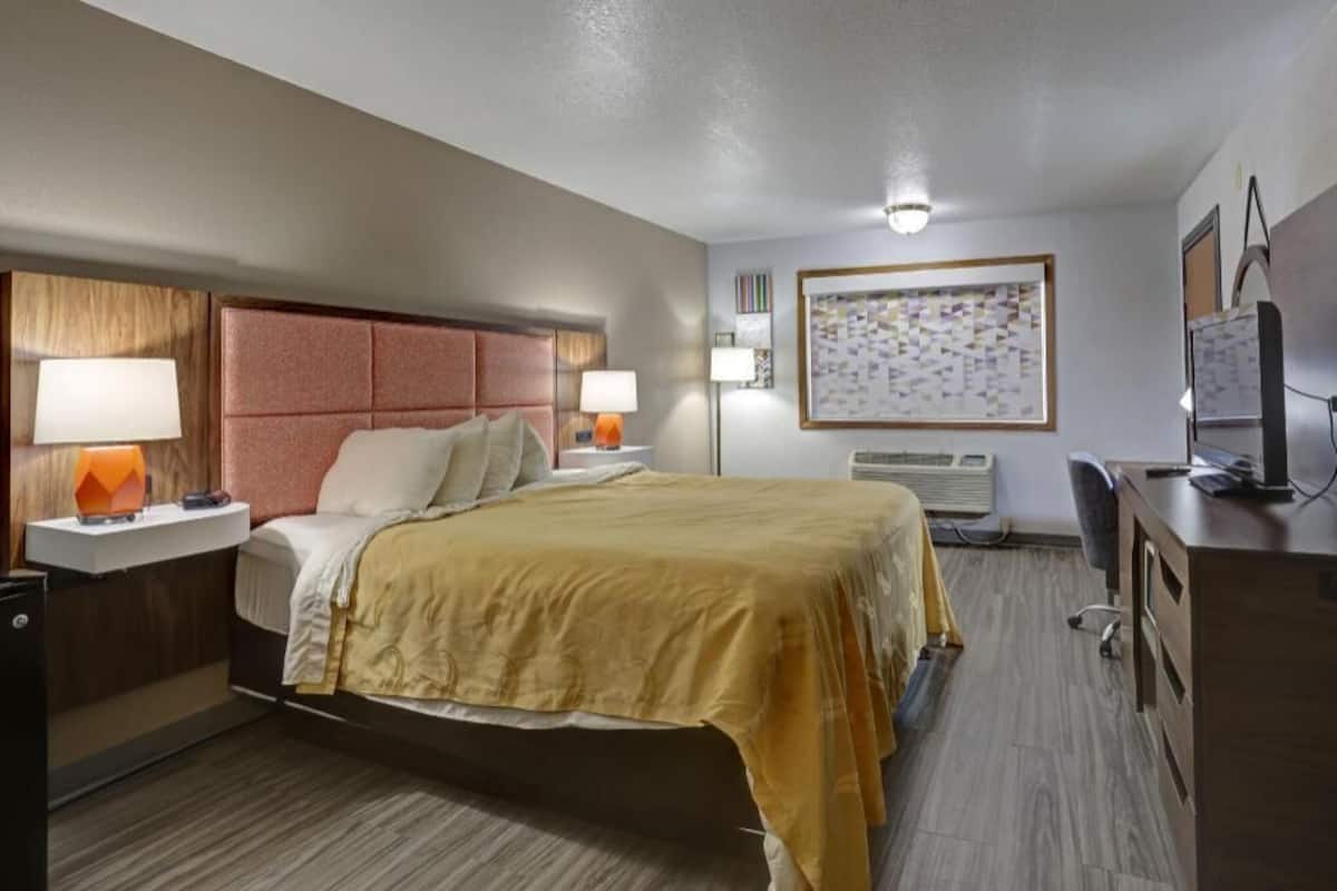 A hotel room with a bed, bedside tables, a desk, a chair, a television, and a wall-mounted air conditioner.
