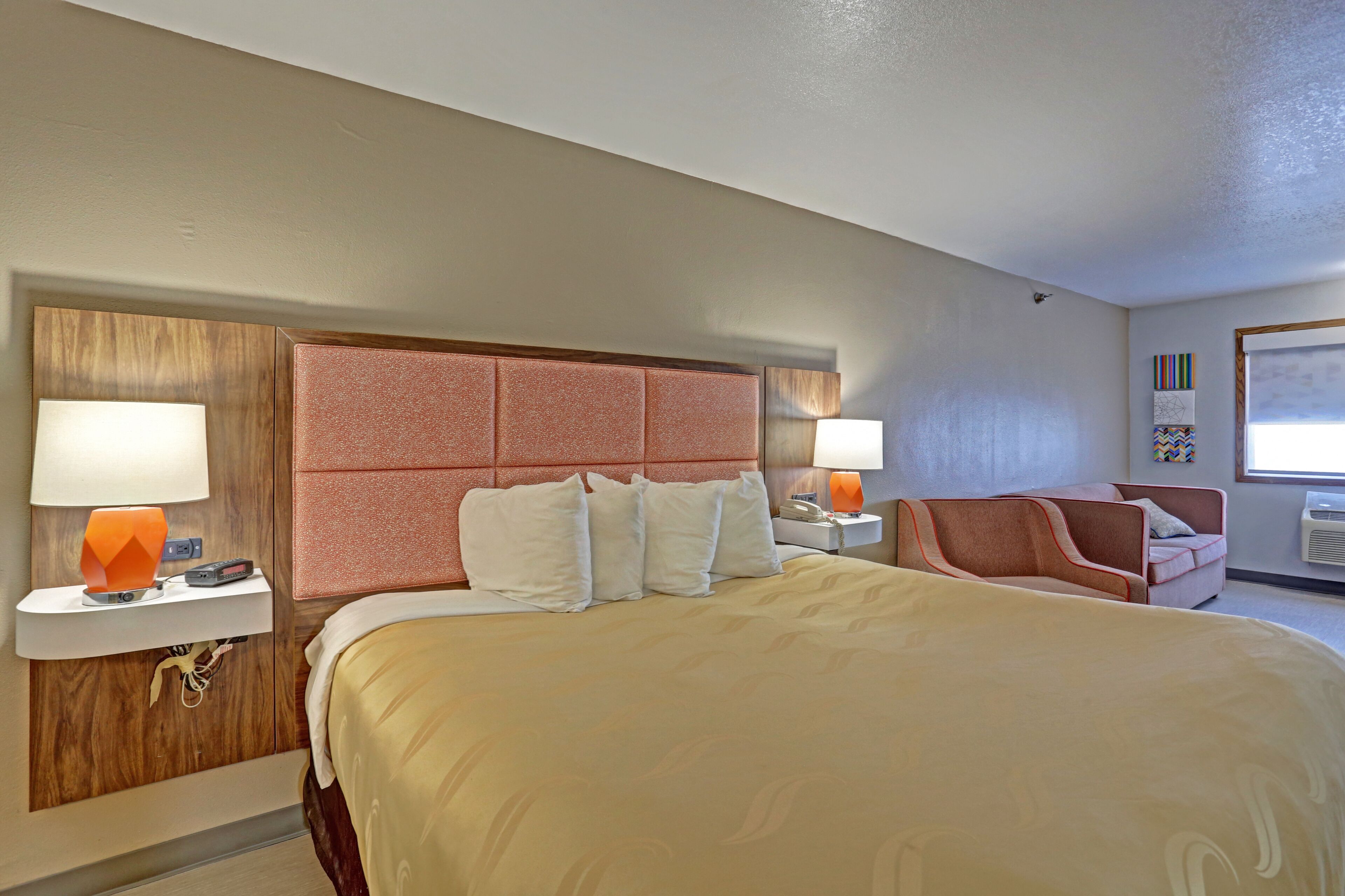 Una habitación de hotel con una cama grande, dos mesitas de noche con lámparas, un cabecero de madera, un sillón rosa y una ventana con persianas.