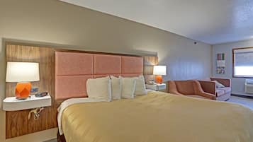 Una habitación de hotel con una cama grande, dos mesitas de noche con lámparas, un cabecero de madera, un sillón rosa y una ventana con persianas.