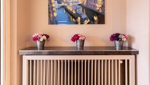 A wall-mounted painting of a cityscape with a river, three flower vases on a console table, and a radiator below.
