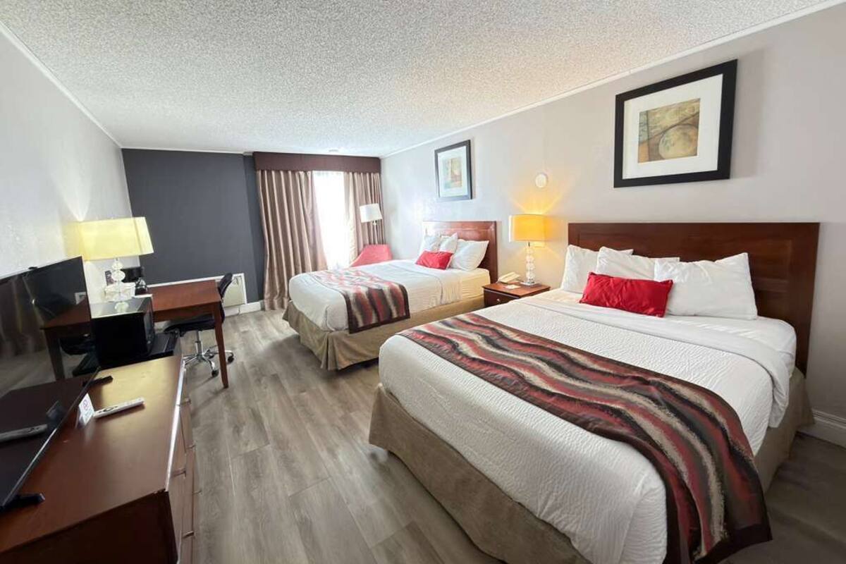 A hotel room with two beds, a desk, a chair, a TV, and a lamp.