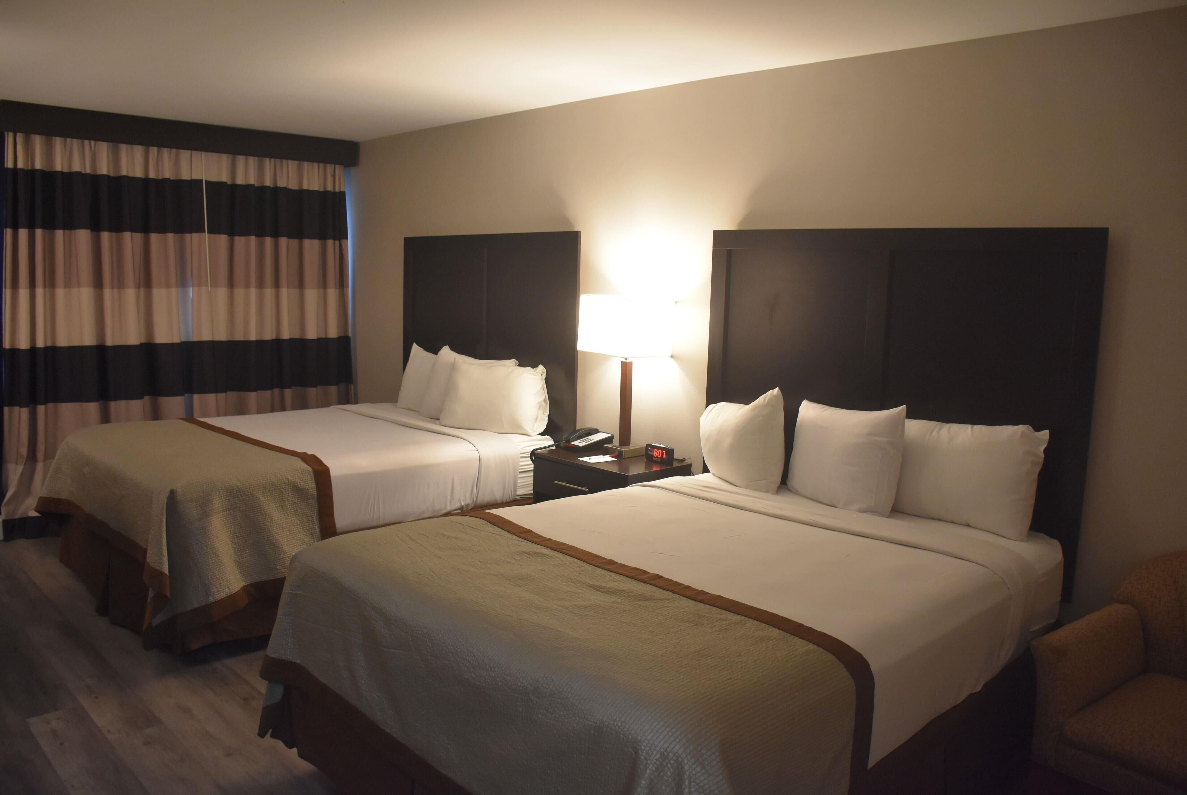 A hotel room with two beds, a nightstand, a lamp, and a chair.