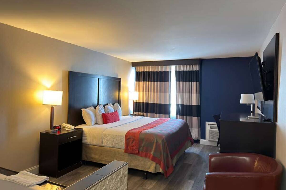 A hotel room with a bed, bedside table, lamp, chair, and a TV.