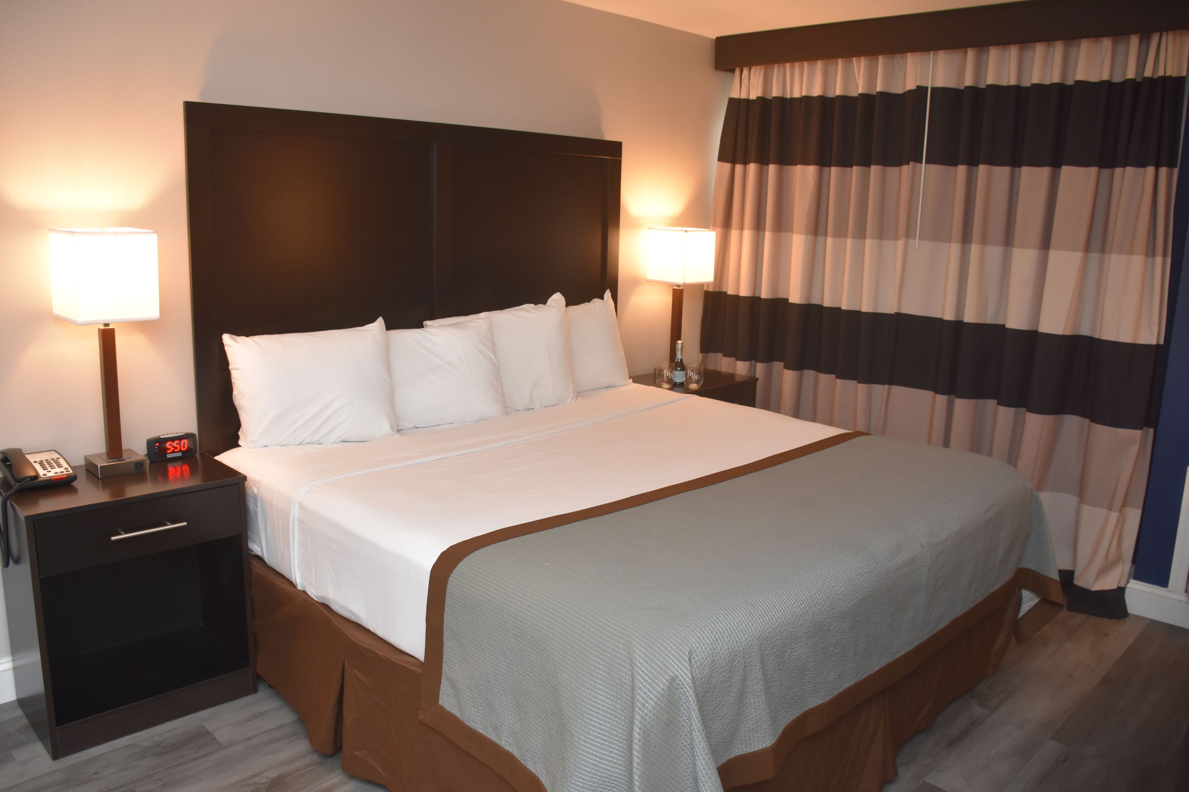 Een hotelkamer met een groot bed, twee nachtkastjes met lampen, een bijzettafeltje met een telefoon en een raam met gestreepte gordijnen.